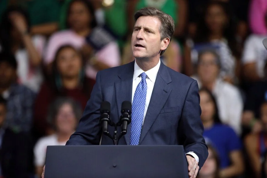 A man in a blue suit and blue polka dot tie speaking at a podium with two microphones, with a blurred crowd of diverse people in the background.