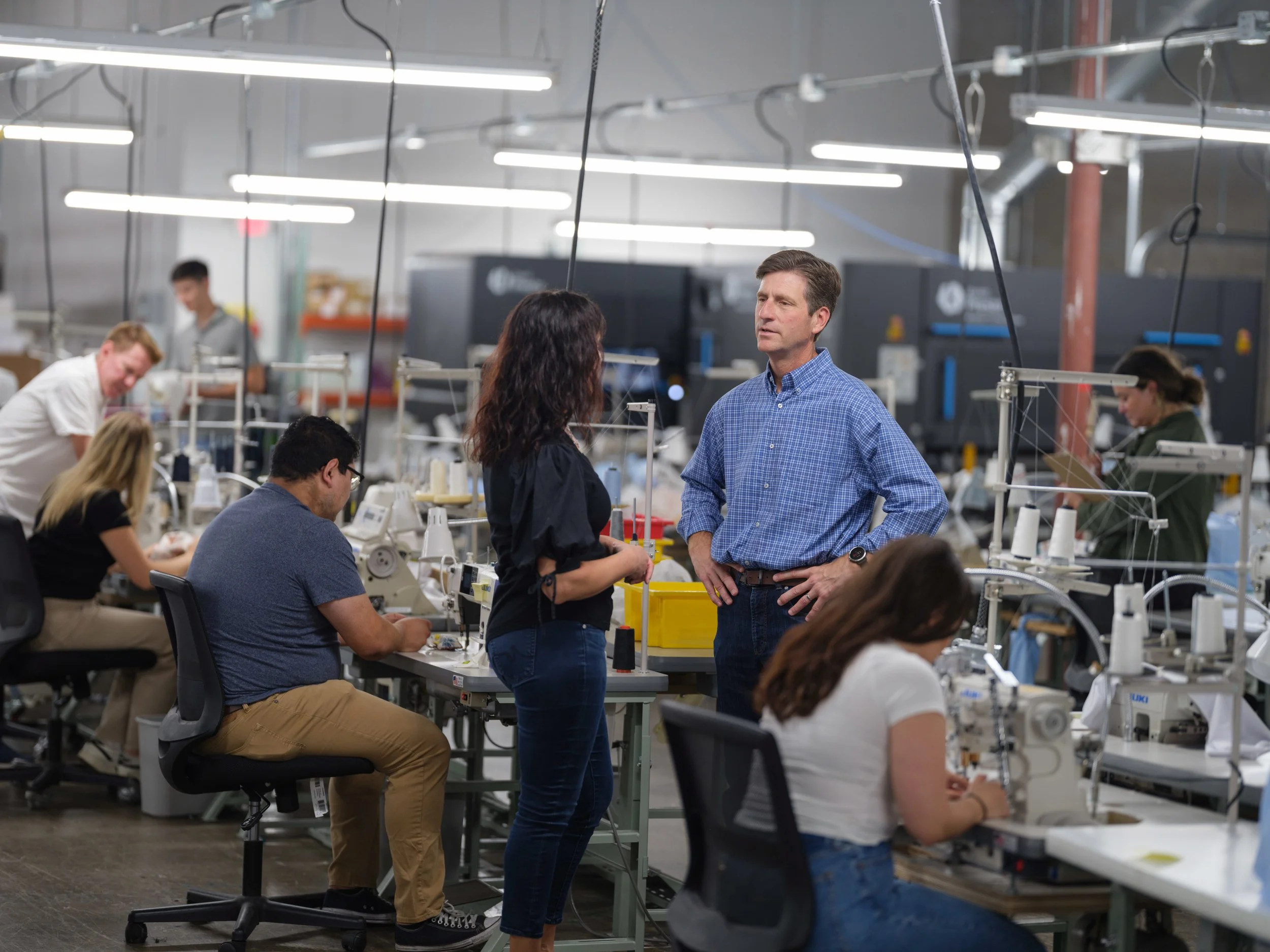 People working on manufacturing or assembly line in a factory setting, with long work tables, sewing machines, and bright overhead lighting.