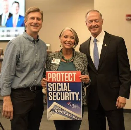 Three people standing together, holding a sign that says 'Protect Social Security.' They are smiling at an indoor event.