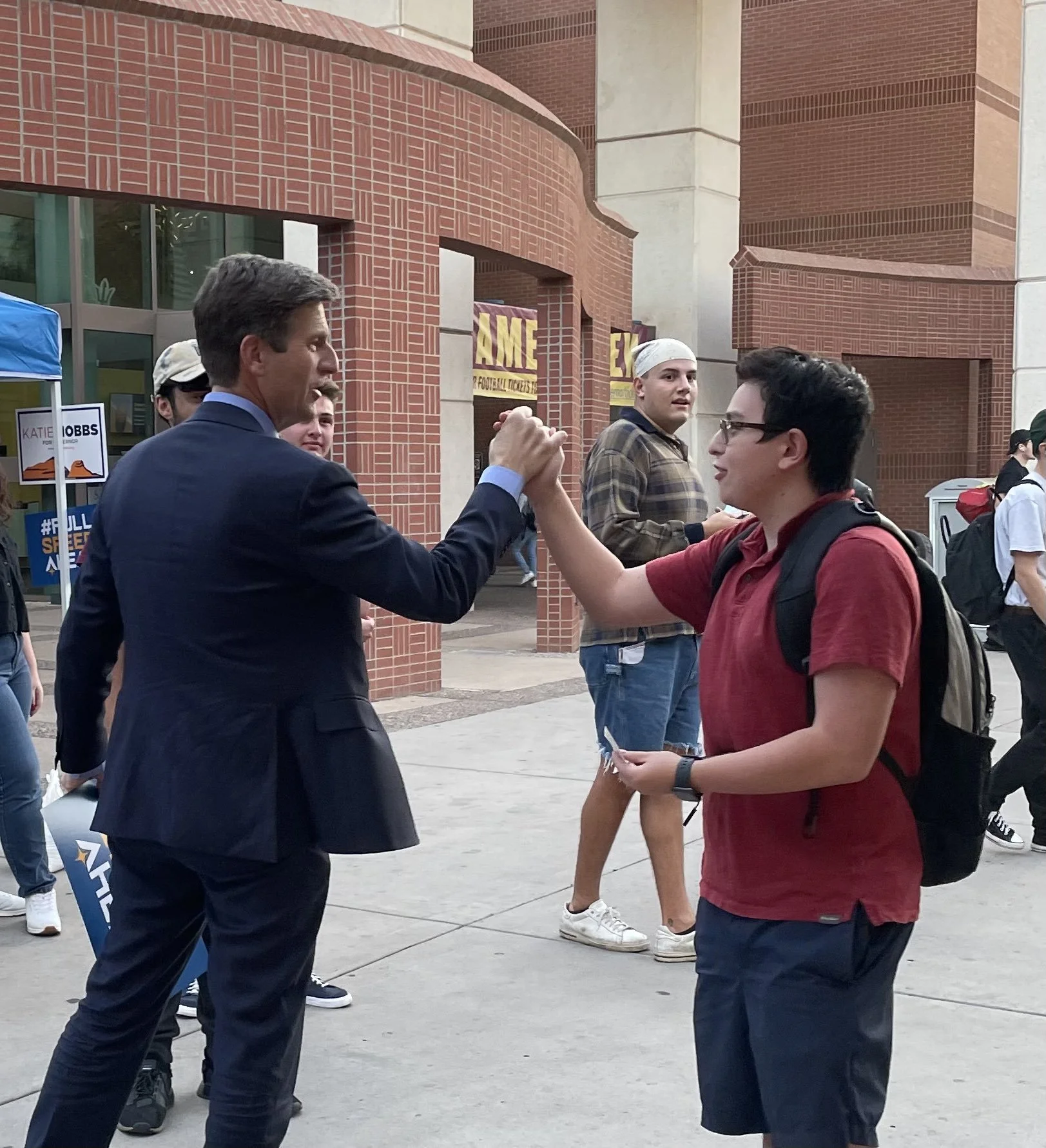 Two men are shaking hands in a social setting outside a brick building. One man is dressed in a suit, and the other is wearing a red polo shirt and glasses. There are other people in the background, some holding signs and wearing backpacks.