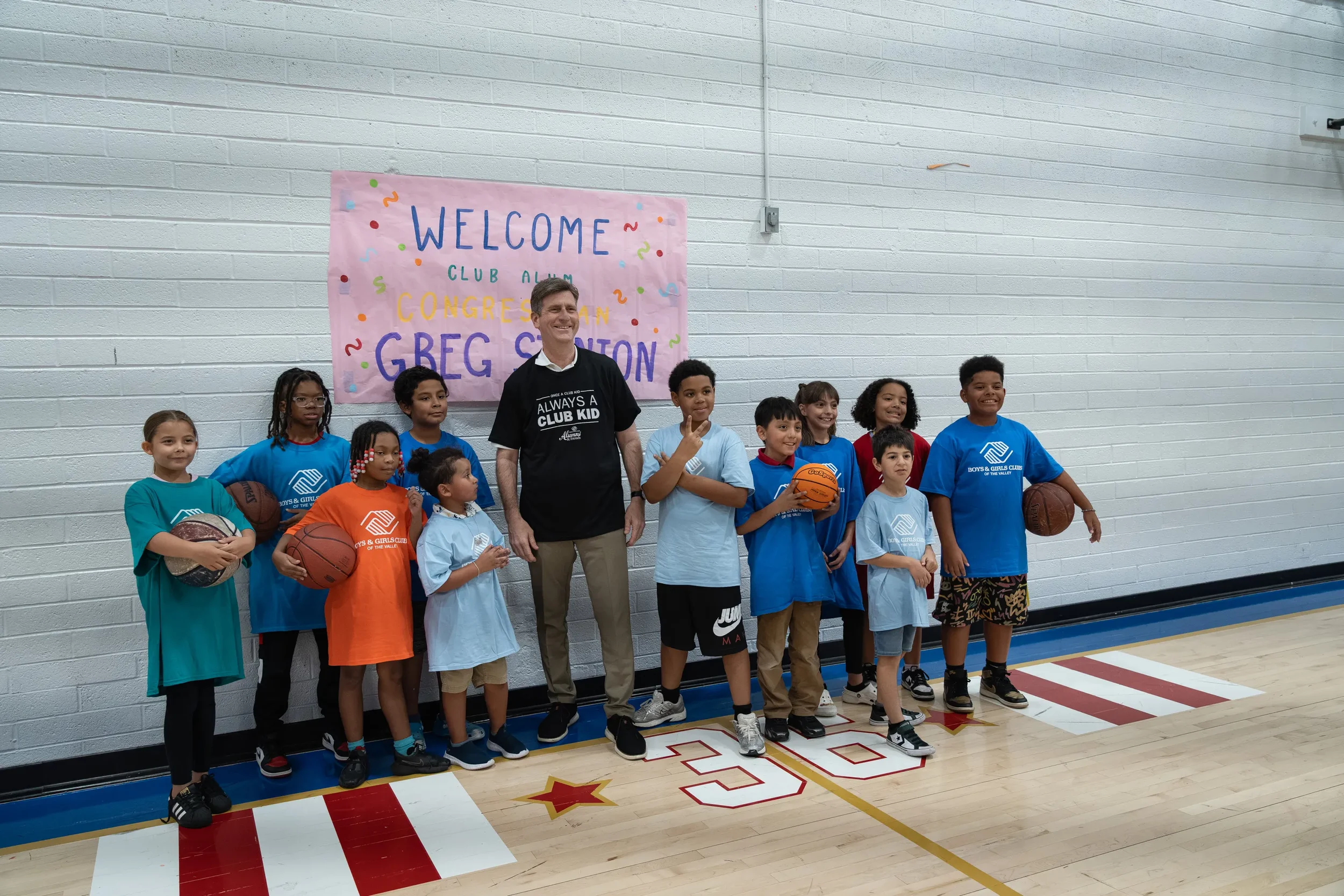 A group of diverse children standing in a gymnasium, holding basketballs, with an adult man in the center, smiling. Behind them, a colorful banner reads 'Welcome Club Alum Congratulation' in front of a white brick wall.