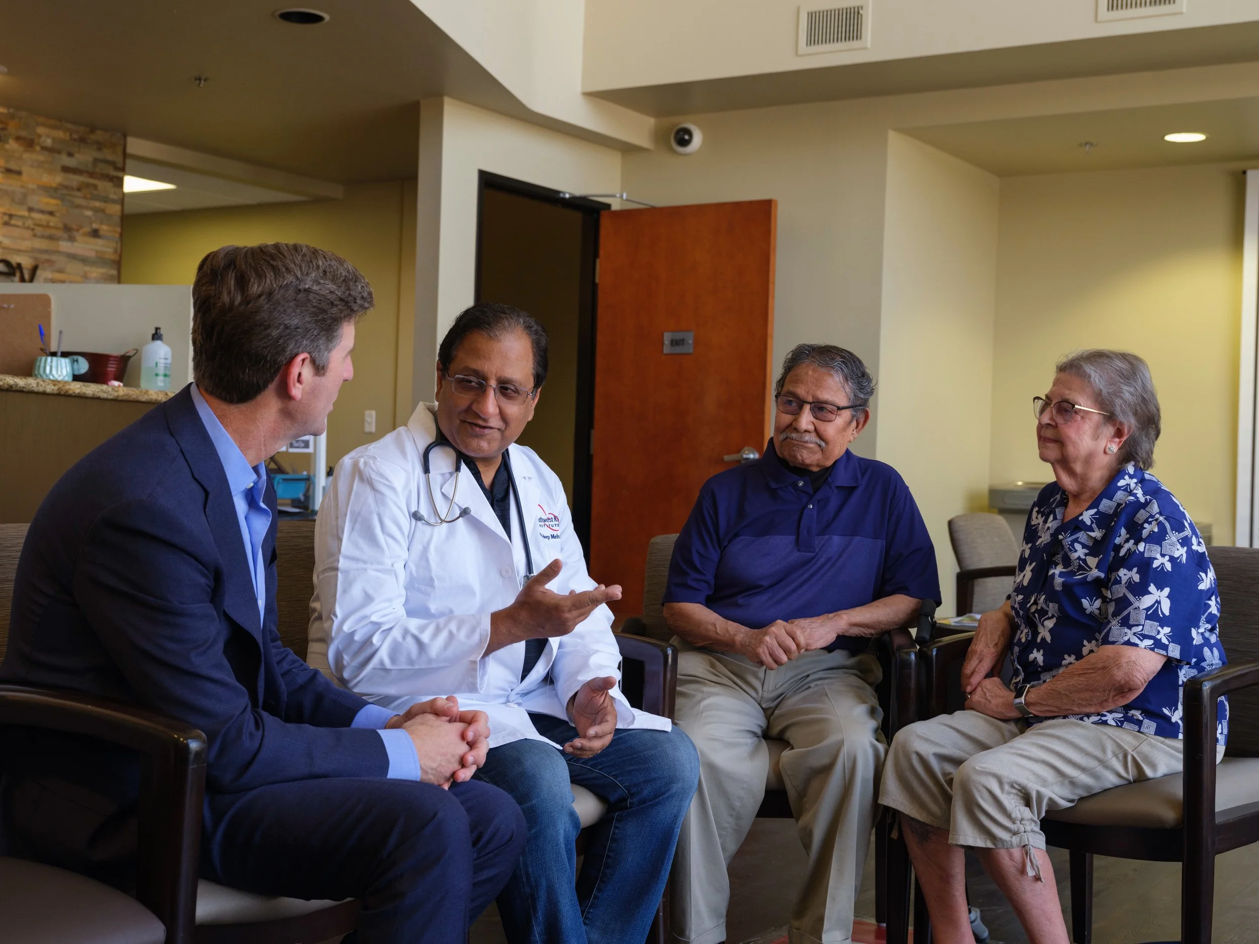 Four adults sitting in a circle engaged in conversation in a room that appears to be a healthcare facility or waiting room.