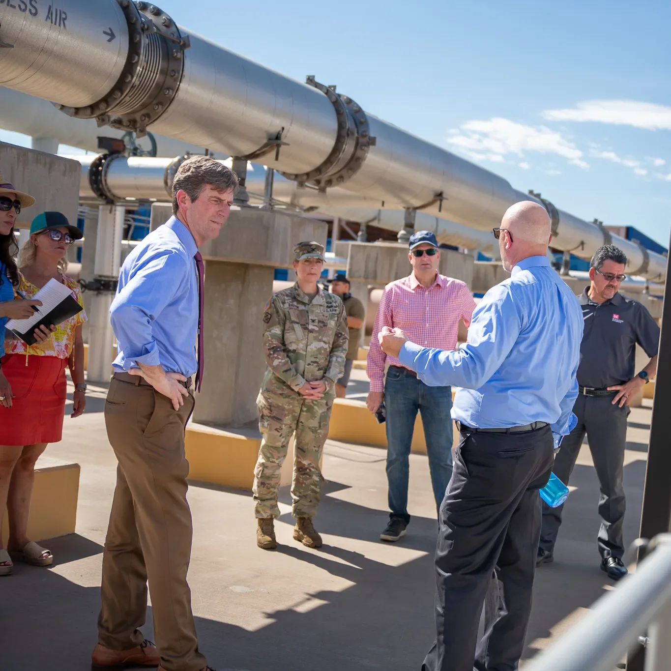 A group of people, including a woman in a colorful shirt, a man in a purple tie, a soldier in uniform, and several men in business casual attire, are gathered outdoors near a large pipeline structure under a bright blue sky with a few clouds, engaged in a discussion or tour.