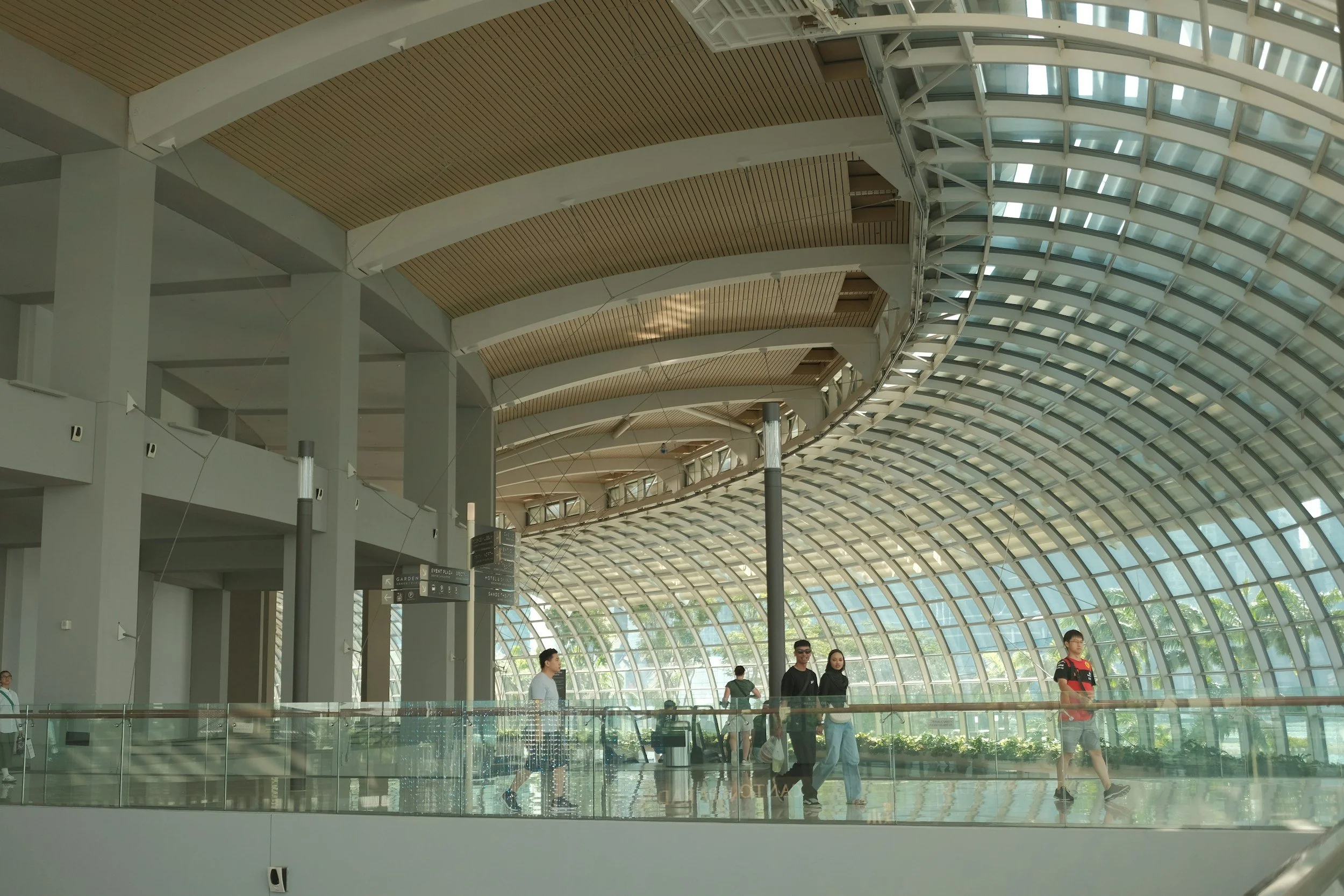 Interior of a modern building with a high glass arched ceiling, multiple people walking around, and directional signs.