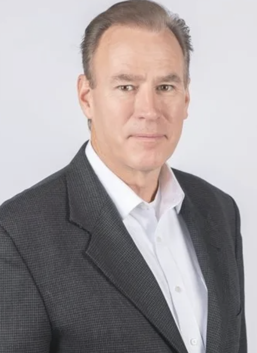 A middle-aged man with light skin, short light brown hair, wearing a dark gray suit and white shirt, against a plain light gray background.