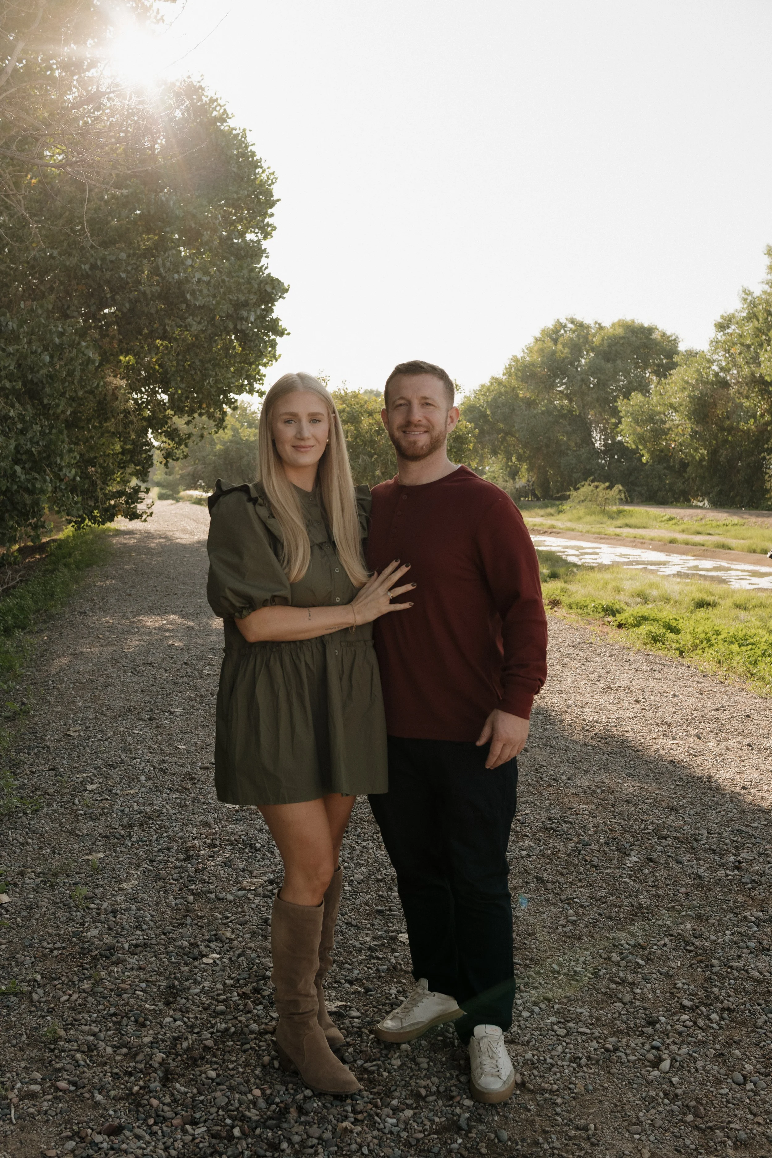 A man and woman smiling and standing together outdoors on a gravel path with trees and sunlight behind them.