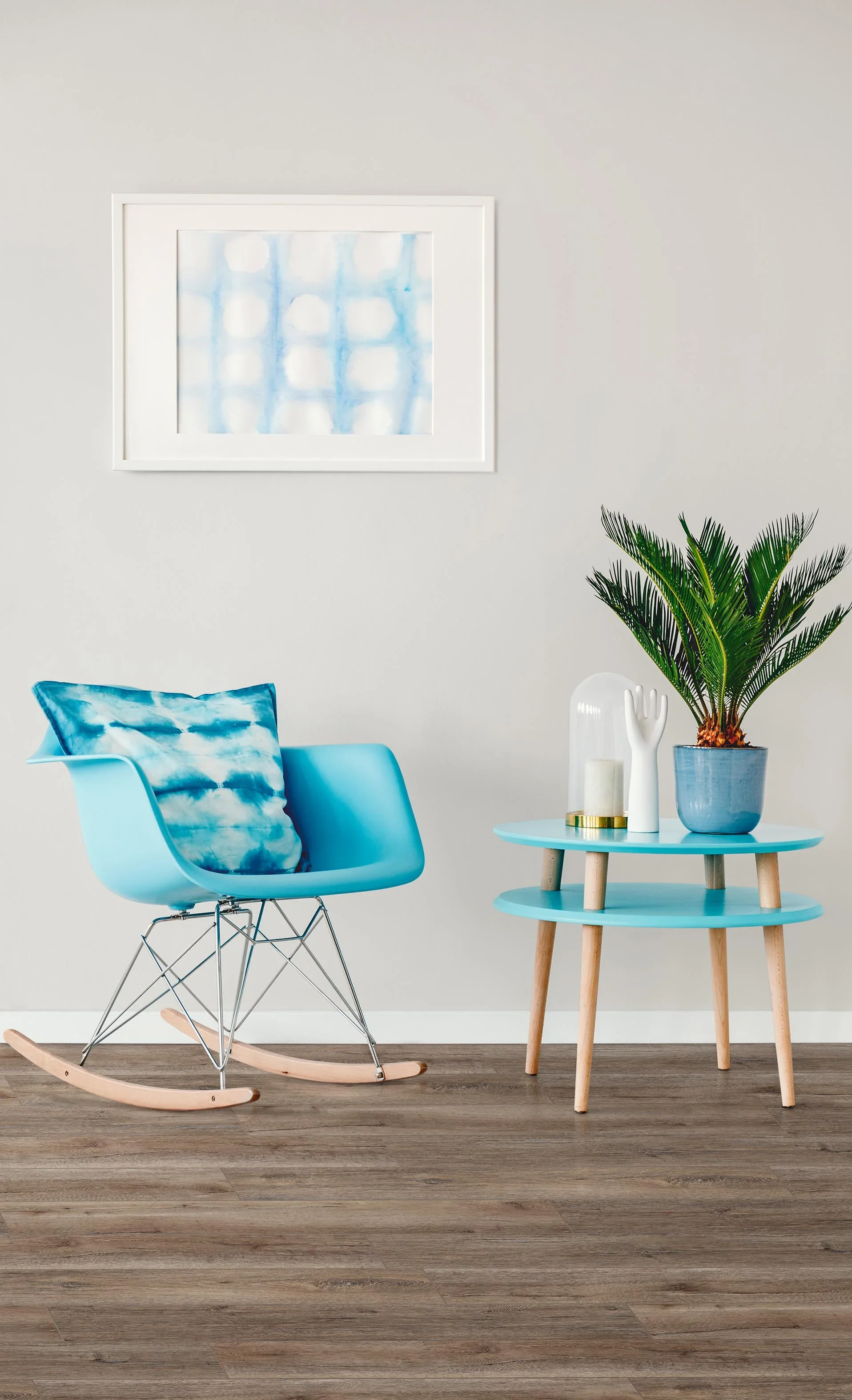 A modern living space with a light gray wall, a white-framed watercolor painting, a bright blue rocking chair with a matching cloud-patterned pillow, a small round table with blue shelves supported by wooden legs, a potted palm plant, and decorative items including a white hand sculpture and candle.