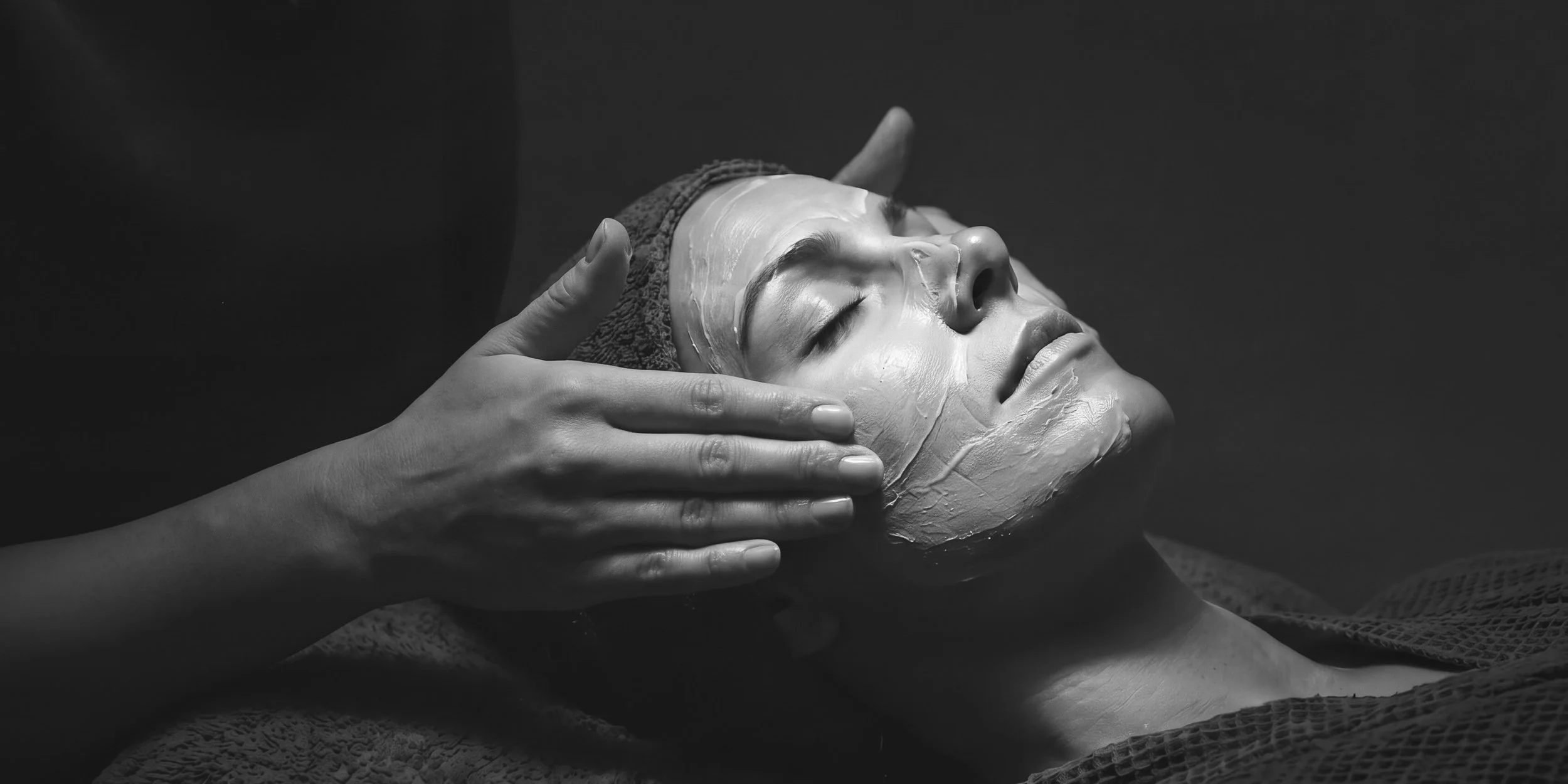 A person receiving a facial treatment with a facial mask applied, lying down with eyes closed, while a therapist gently touches the person's face.