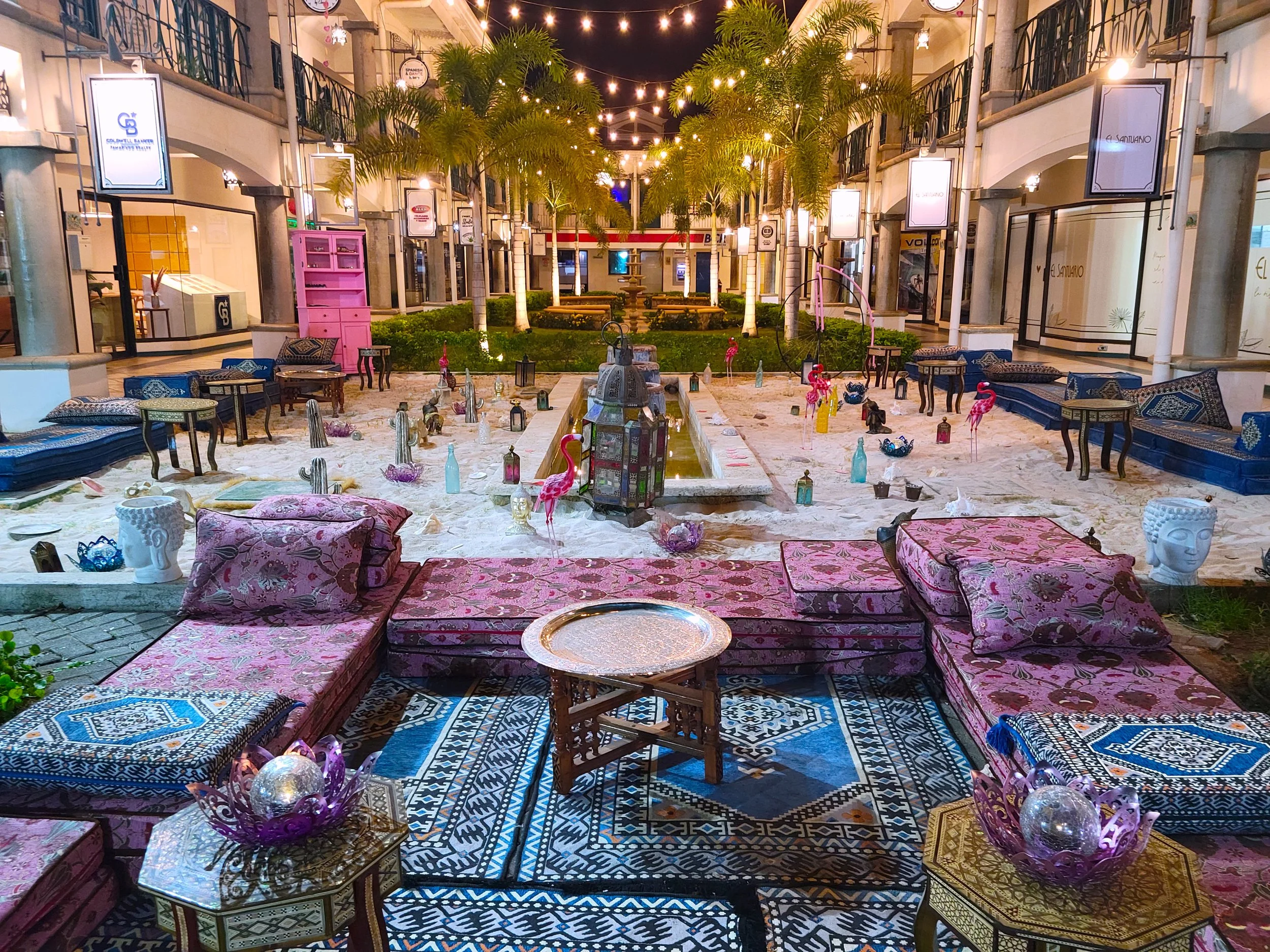 Outdoor courtyard at night with string lights, palm trees, and a fountain in the center, decorated with pink flamingo statues, lanterns, and cushions on patterned rugs and sofas, surrounded by storefronts with signs.