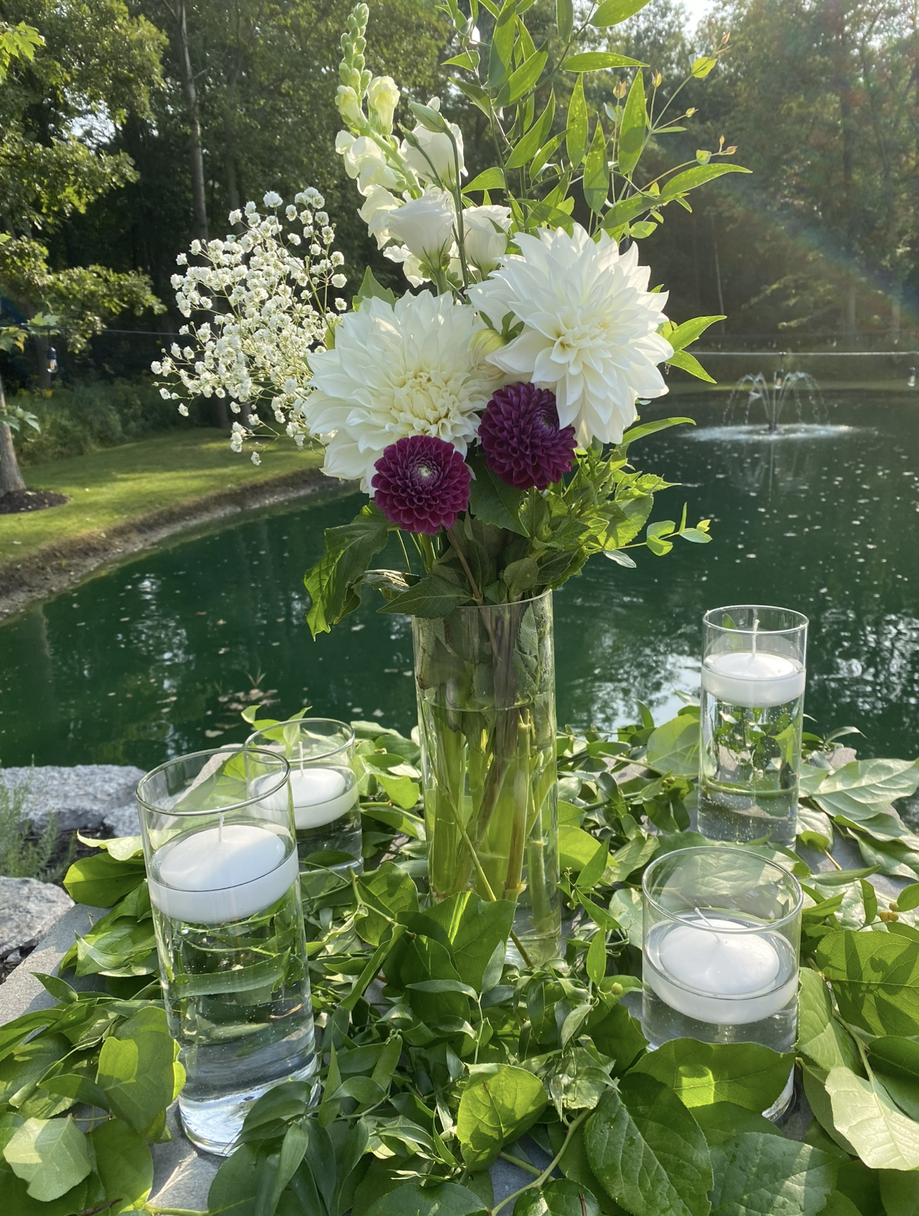 A floral centerpiece featuring white and purple flowers in a tall glass vase, surrounded by five smaller glass candles filled with water and floating candles, set outdoors near a pond with trees and a fountain.