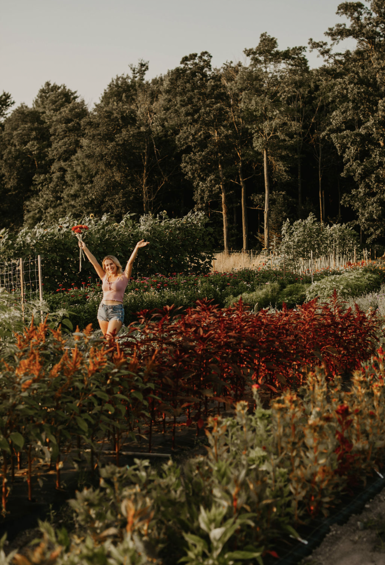 A woman with blonde hair wearing a pink sleeveless top and denim shorts standing in a flower garden, holding a bouquet of flowers in one hand and raising her other arm in celebration.