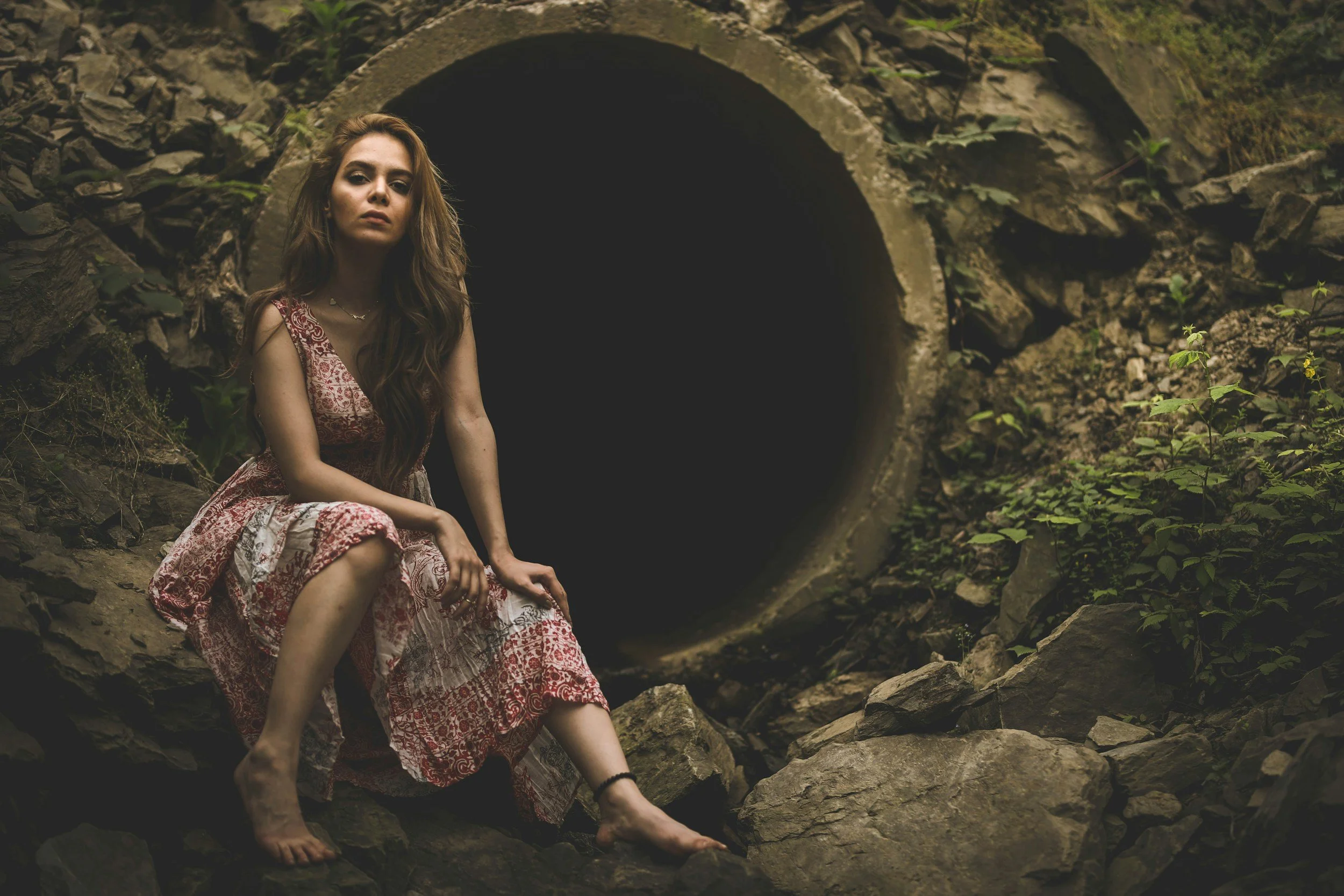 A woman with long brown hair sitting on rocks near a large dark tunnel opening in a rocky, green environment.
