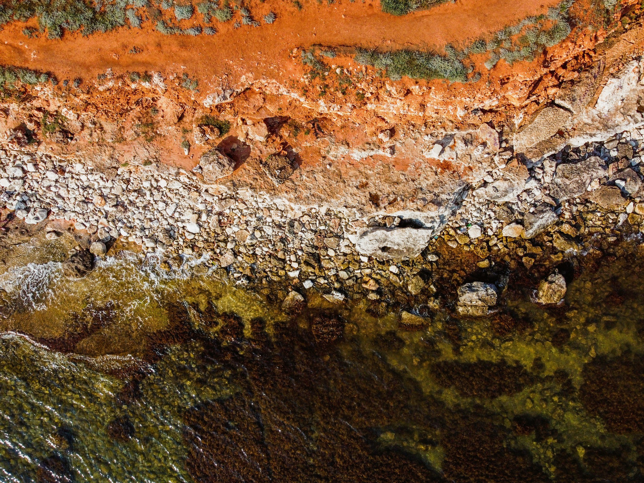 Aerial view of a red-orange rocky cliff with sparse vegetation and a small rocky beach area at the bottom, next to water.