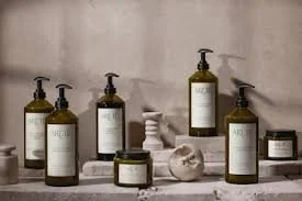 Collection of Arete hair care in amber and clear glass bottles and jars with black pump dispensers and labels, arranged on a gray surface against a plain wall.