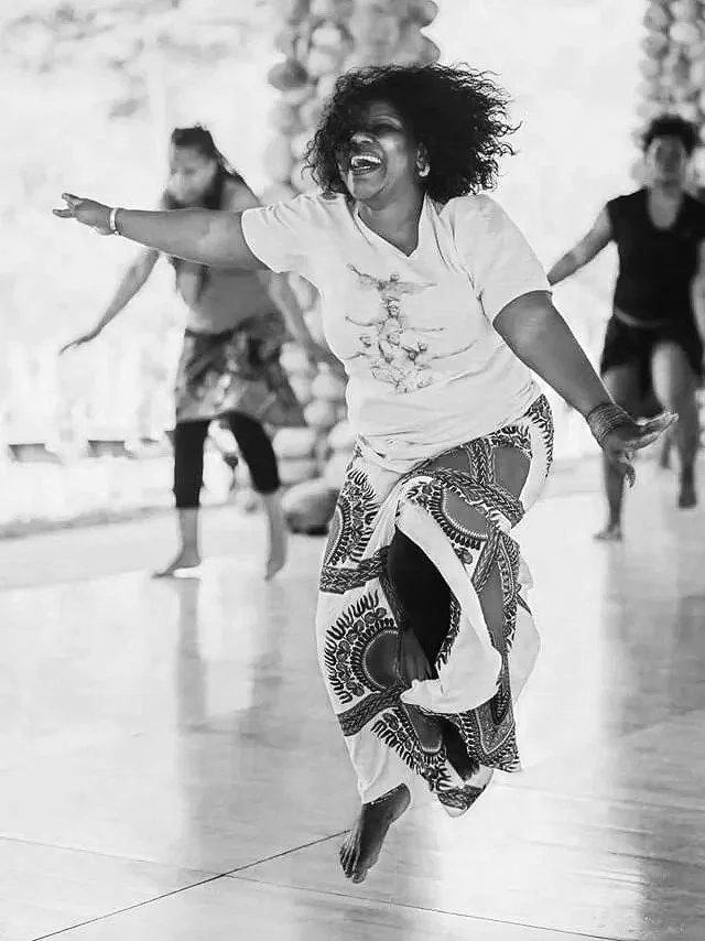 A woman with curly hair dancing joyfully in a spacious room, wearing a white t-shirt and patterned skirt. She appears to be leading or participating in a dance class or cultural activity, with other women dancing in the background.