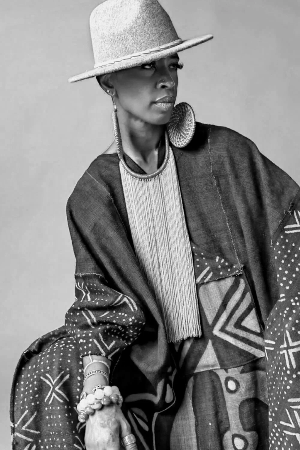 Black and white portrait of a woman wearing a large hat, earrings, a beaded necklace, and a patterned, loose-fitting outfit with various geometric designs.