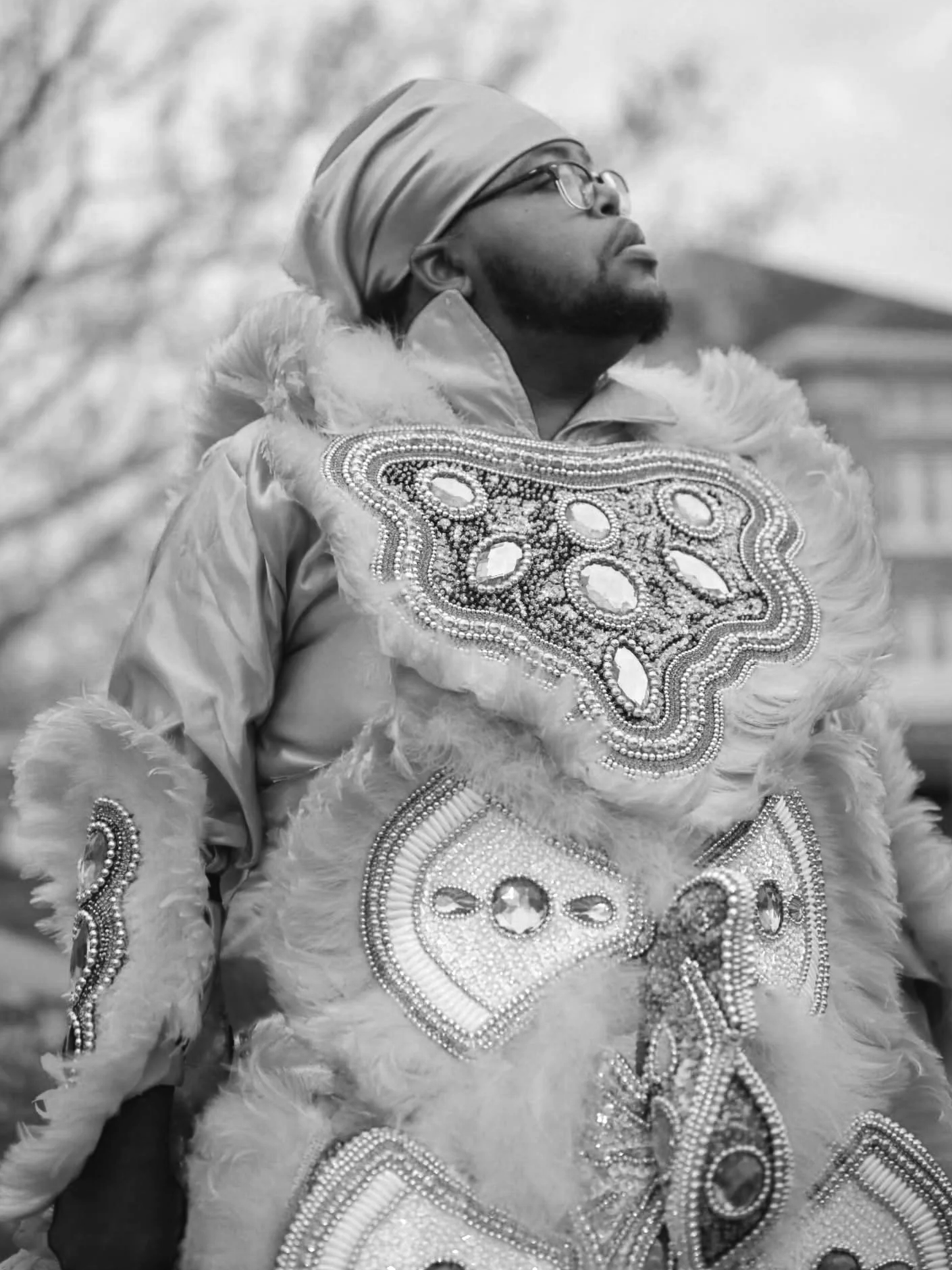 A man in elaborate costume with beaded decorations and feathers, looking upwards, wearing glasses and a turban, in black and white photography.