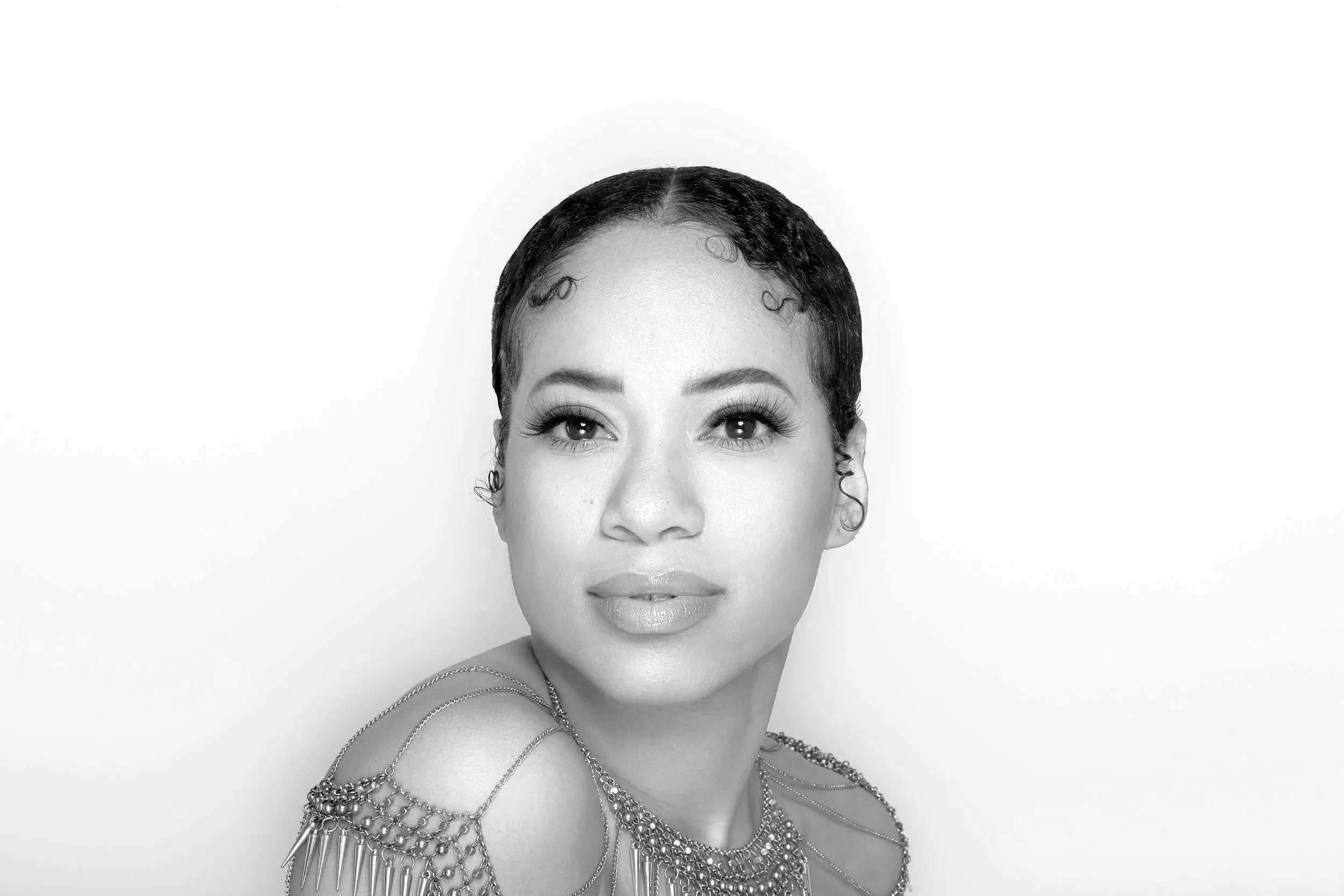 Black and white portrait of a woman with short, curly hair, wearing jewelry including necklaces and earrings, looking directly at the camera with a confident expression.