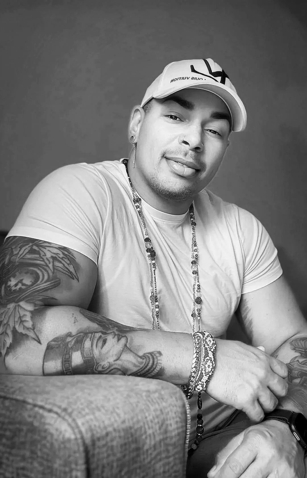A young man with tattoos on his arms wearing a white T-shirt, a white cap, and jewelry, smiling at the camera in a black and white photo.