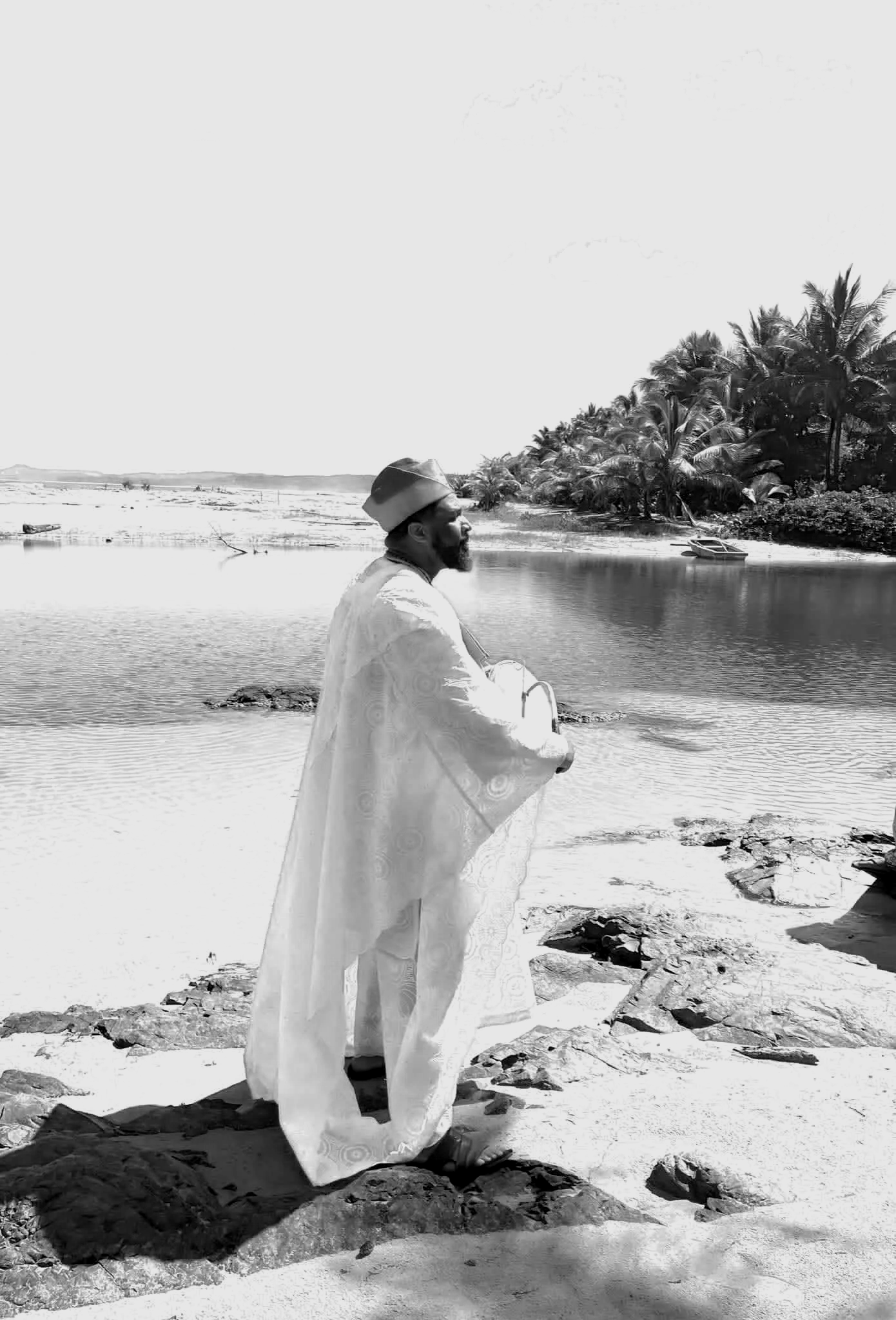 A man dressed in traditional clothing, a white robe, and a hat, standing on a rocky shore near the water, with a background of palm trees and a boat on the beach, in a black-and-white photo.