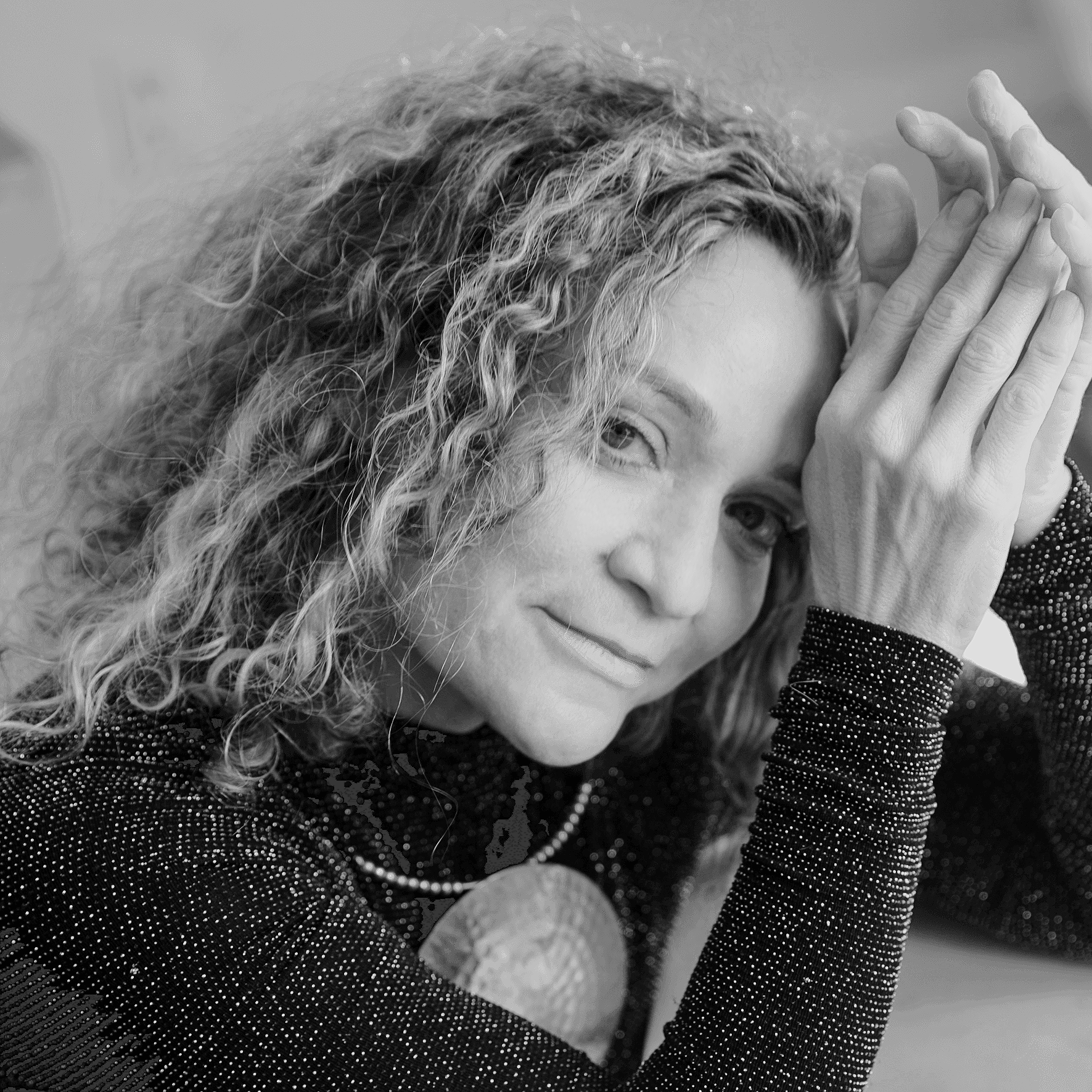 A black and white photo of a woman with curly hair, her hands clasped together near her forehead, smiling softly.