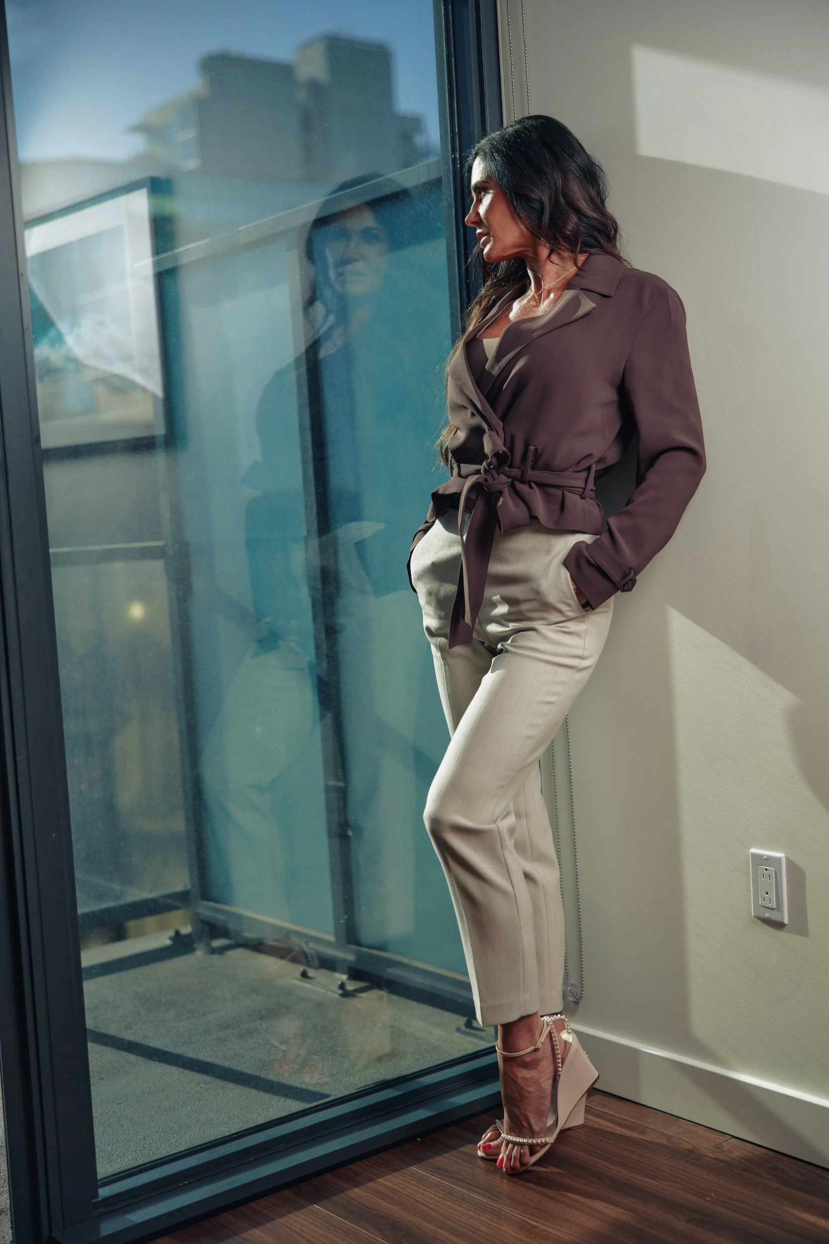 A woman wearing beige pants, a brown blazer, and high-heeled shoes stands by a large glass window, looking outside. Her reflection is visible in the glass, and sunlight casts shadows on the wall. The room has wooden flooring and beige walls.