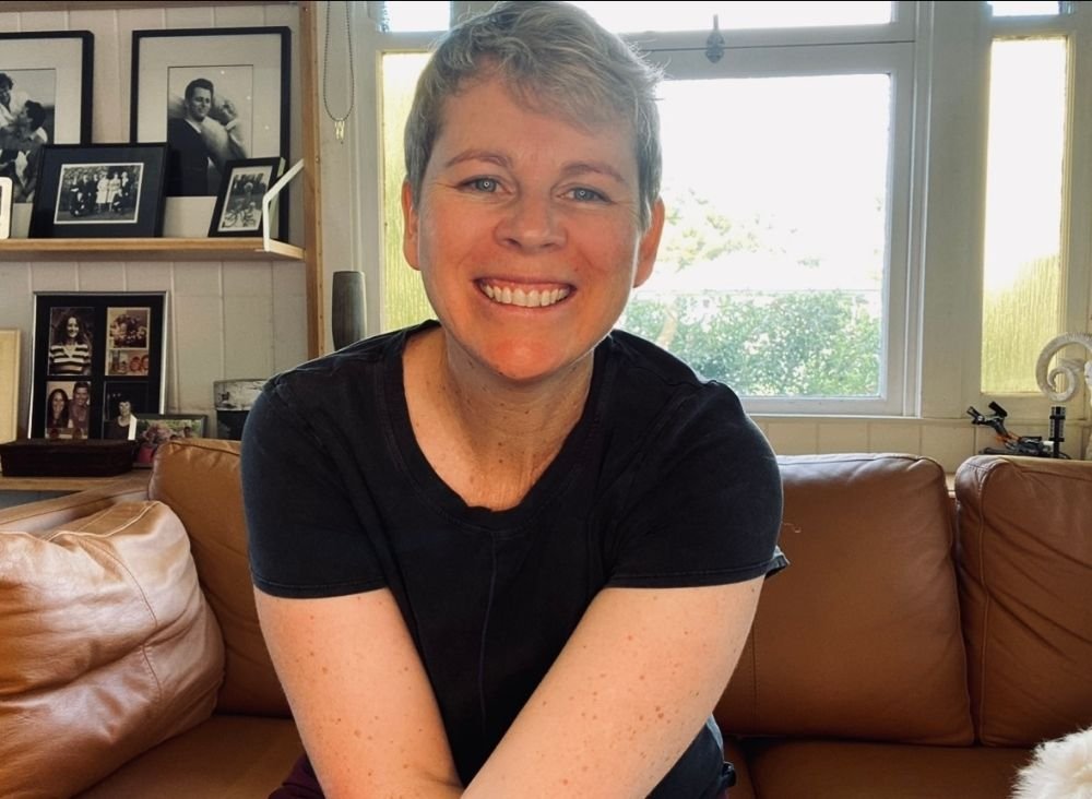 Rachel, pelvic floor exercise physiologist sitting on a couch looking at the camera smiling with a window behind her