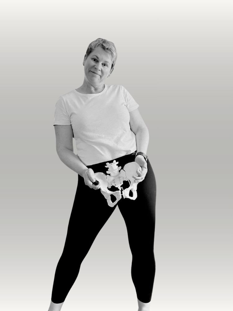 Rachel standing with her hips to her left holding a model pelvis infront of her pelvis. She has balck tights on highlighting the white pelvis and a quirky look on her face.