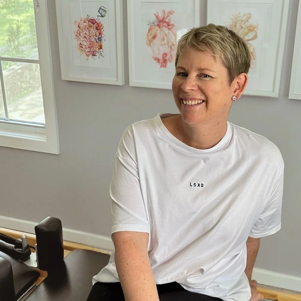 Rachel sitting in her clinic looking at the camera smiling. She is wearing a white t-shirt