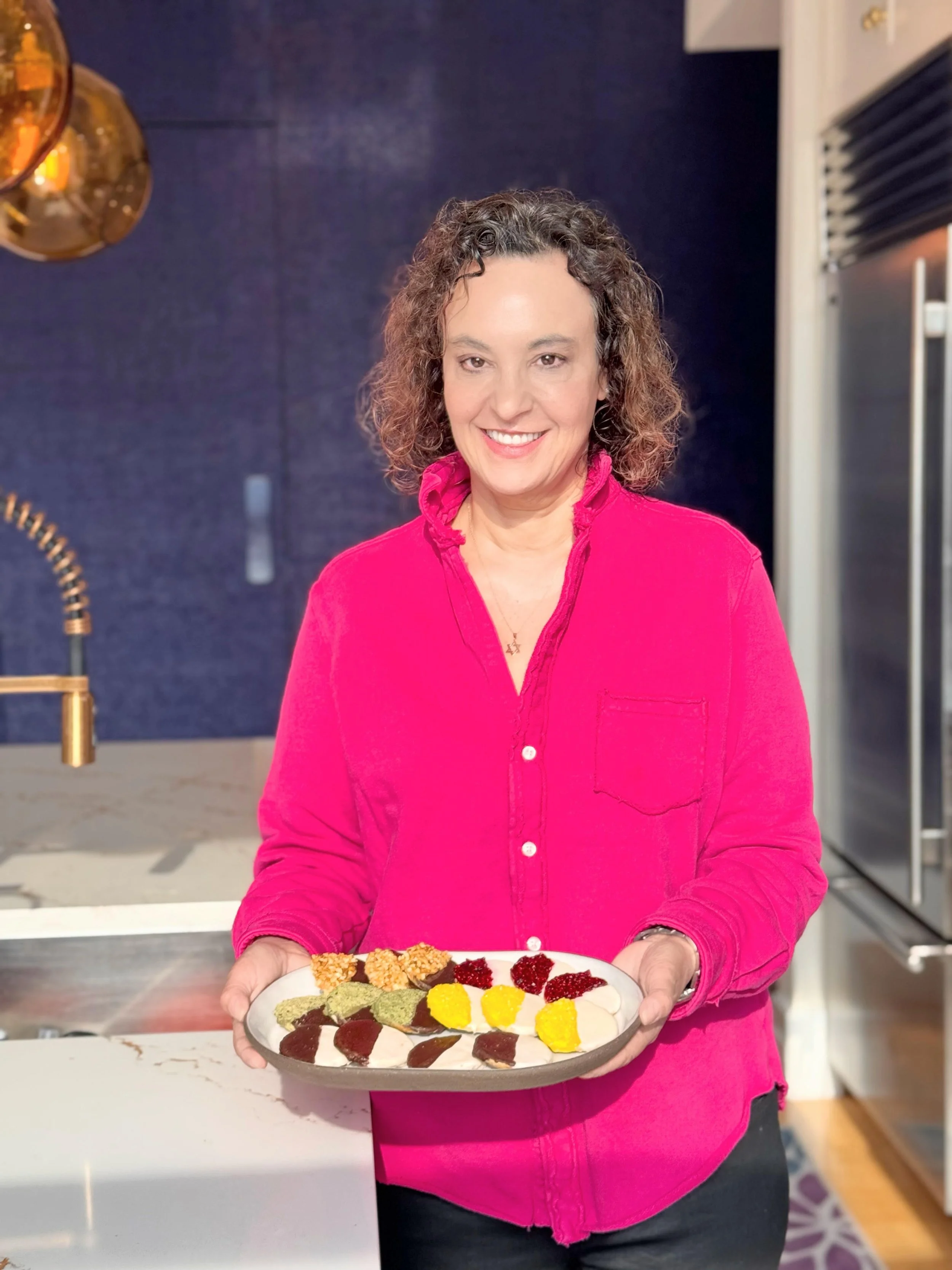 A woman with curly hair wearing a bright pink shirt holding a dessert platter with various colorful desserts in a kitchen.