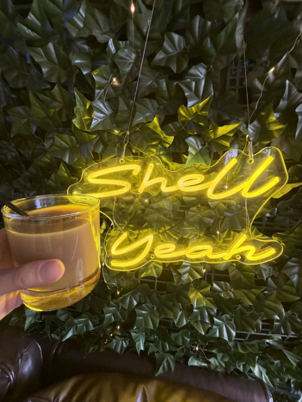 A person's hand holding a glass of a brown beverage in front of a illuminated yellow neon sign that says "Shell Yeah" with green leafy decor in the background.