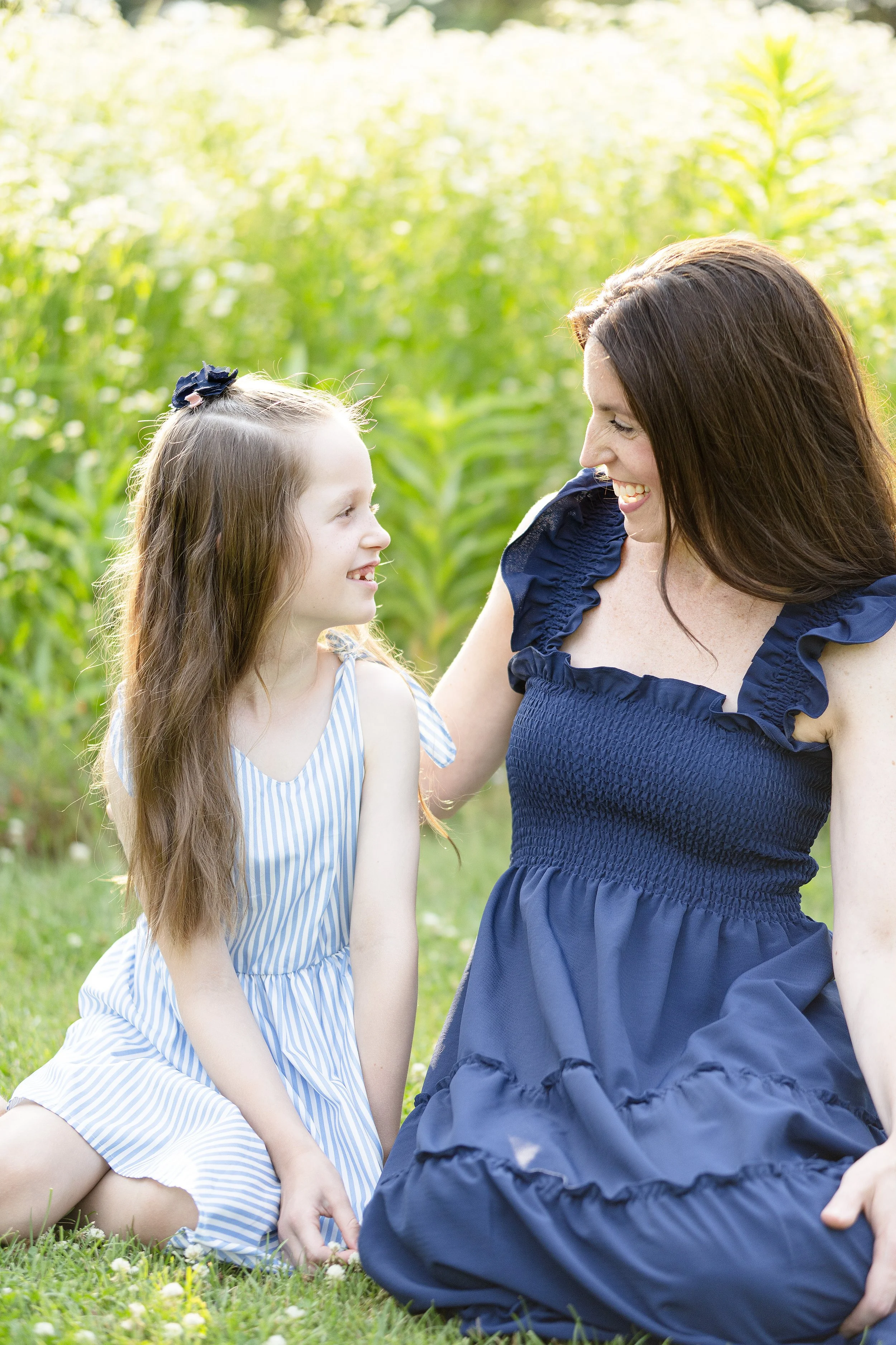 A woman and a young girl sitting on the grass in a lush green field, smiling and looking at each other.