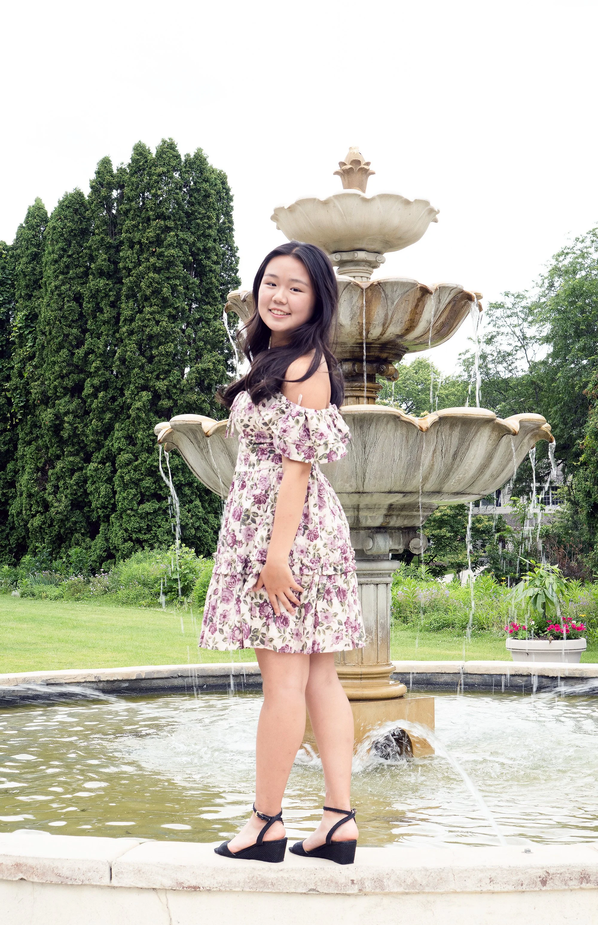 Young woman in a floral dress and black wedge shoes standing by a multi-tiered fountain in a park with trees and flowers.