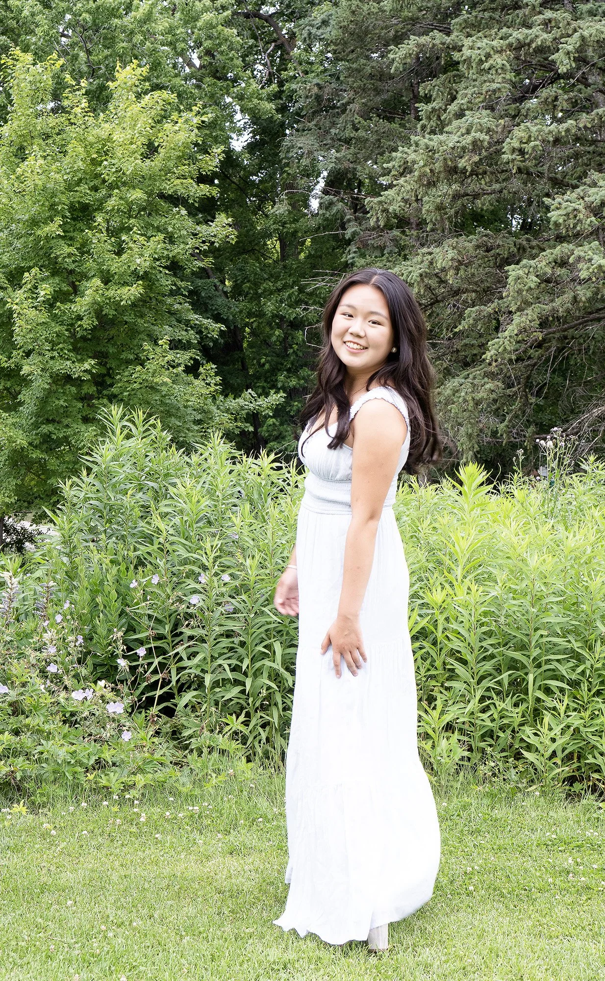 A young woman in a white dress standing in a lush green garden with tall trees and flowering plants, smiling at the camera.