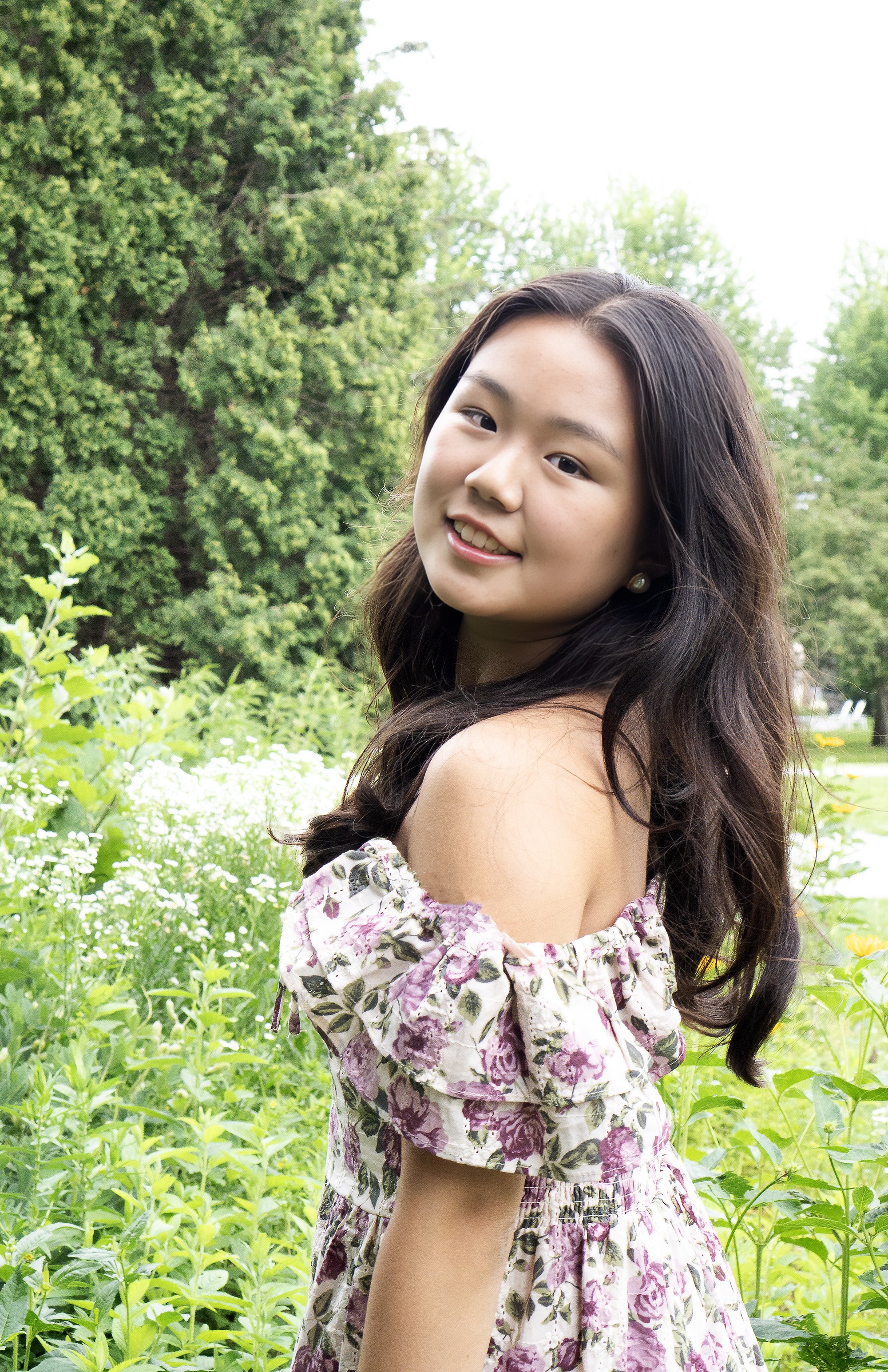 A young woman with long dark hair and pearl earrings smiling outdoors among green trees and flowering plants.