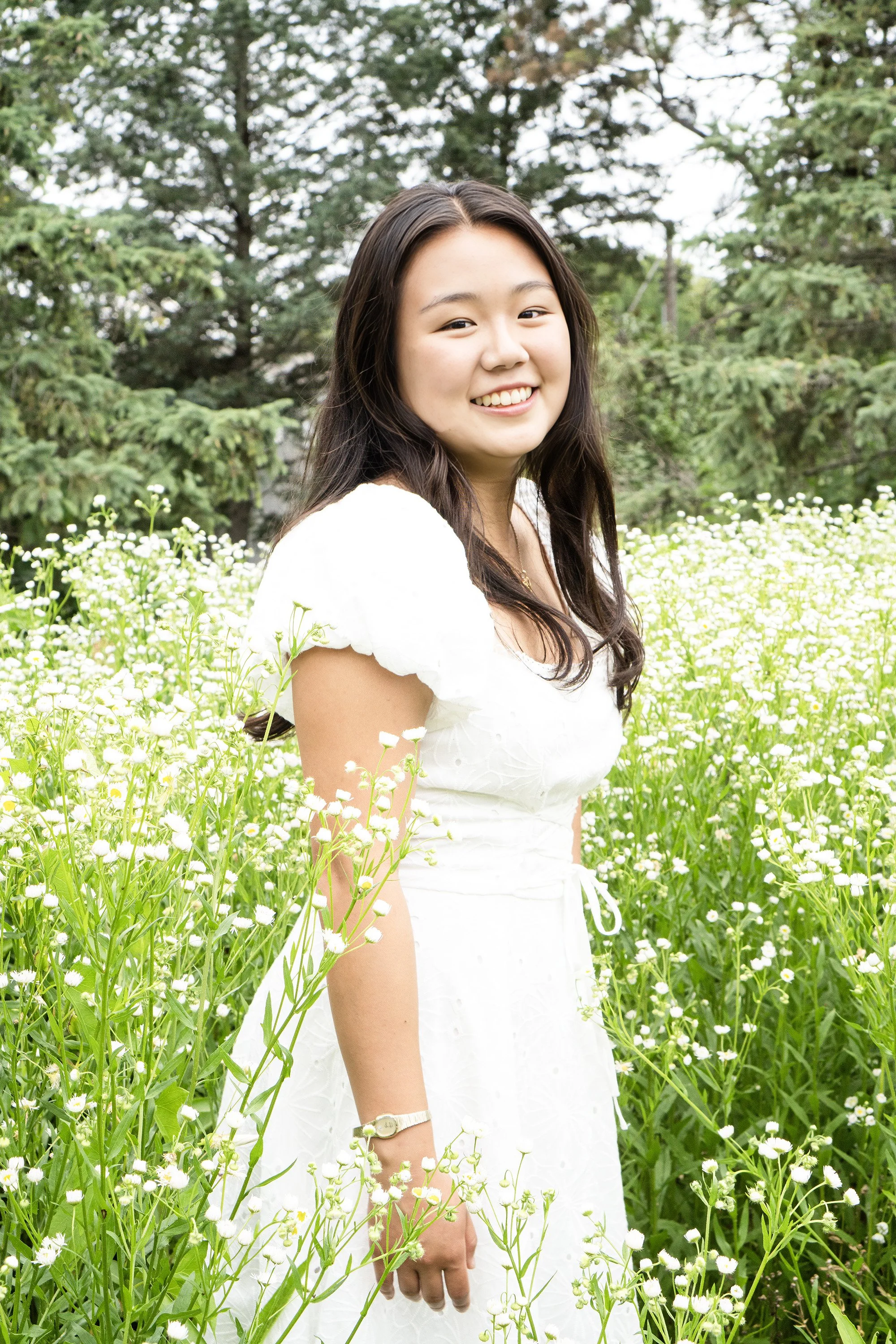 A young woman with long dark hair smiling in a white dress, standing among white daisies in a field with trees in the background.