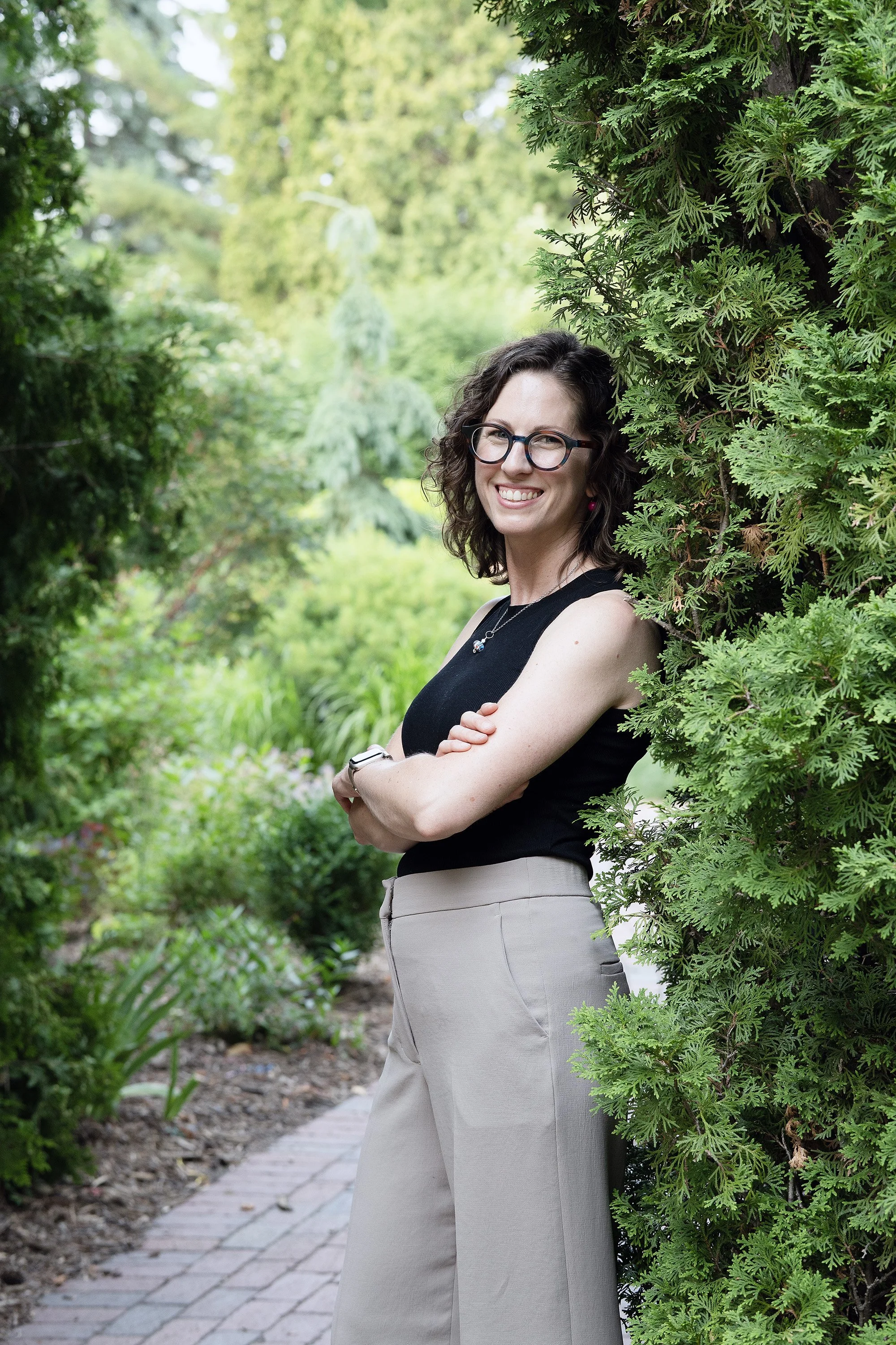 A woman with curly dark hair, wearing glasses, a black sleeveless top, and beige high-waisted pants, smiling with crossed arms, standing beside a lush green bush on a garden path.