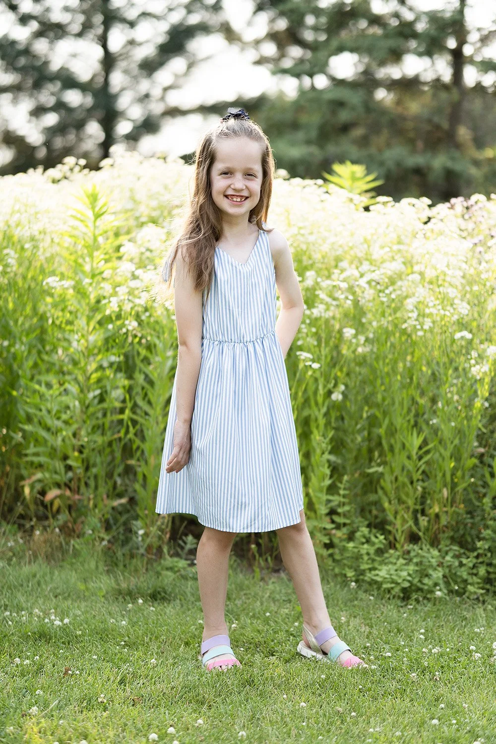 Young girl with long brown hair, wearing a sleeveless blue and white striped dress and pink sandals, standing on grass in front of yellow and white flowers and trees, smiling at the camera.
