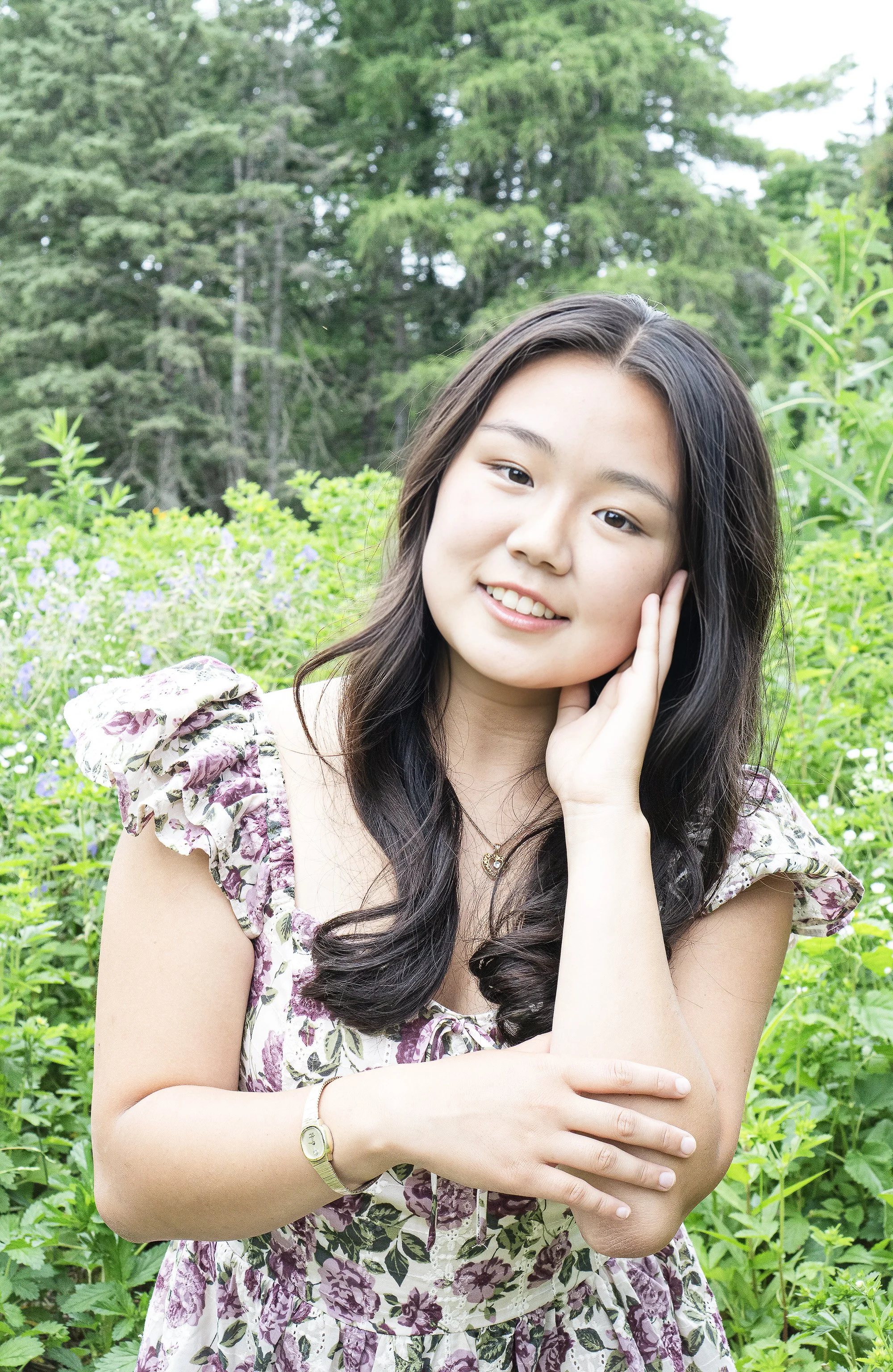 A young woman with long dark hair and a floral dress posing outdoors among greenery, smiling gently with one hand touching her face.