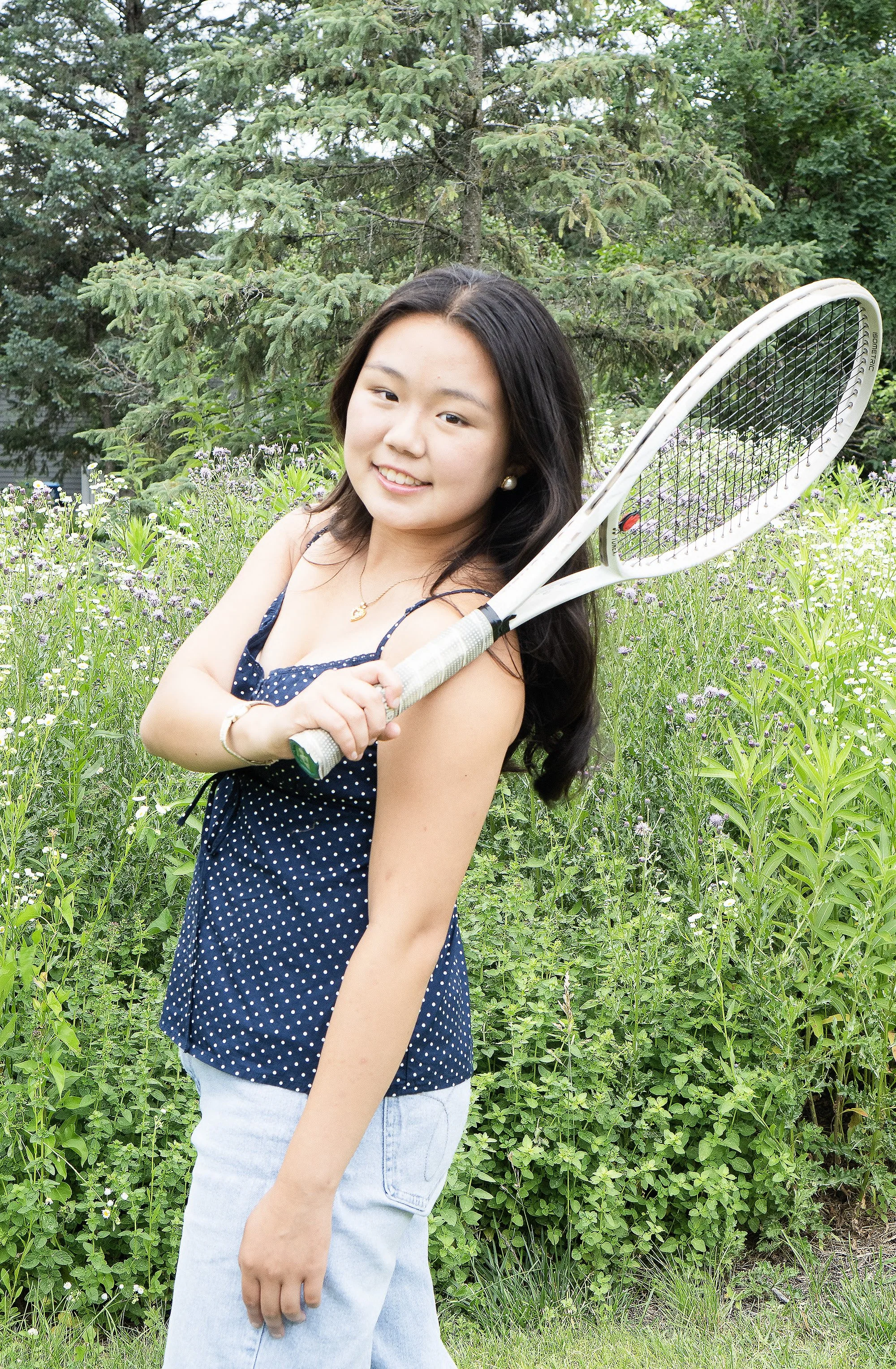 A smiling young woman with long dark hair, wearing a navy blue polka dot sleeveless top and light blue jeans, is standing outdoors in a green field with wildflowers and tall trees in the background, holding a tennis racket.