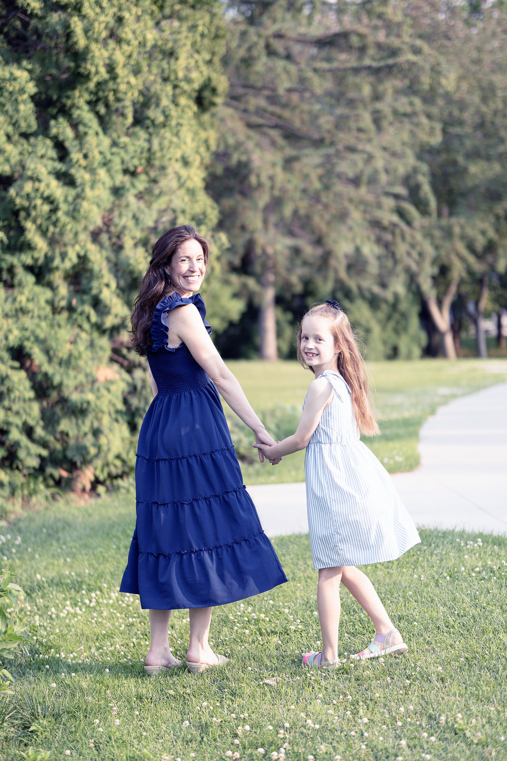 A woman and a girl holding hands in a park, smiling, with trees and grass in the background.