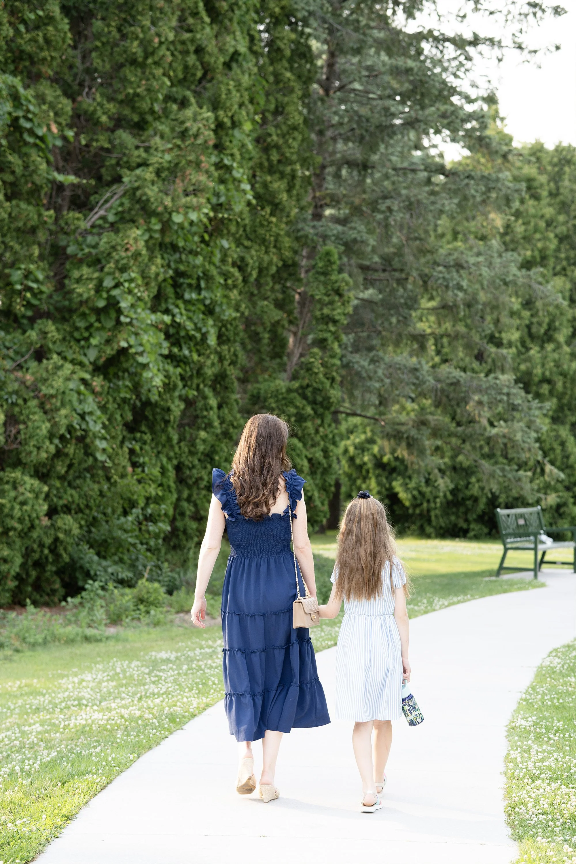 A woman and a girl walking hand-in-hand down a paved path in a park surrounded by lush green grass and trees. The woman wears a blue dress and carries a beige purse, while the girl wears a white dress and carries a patterned bag.