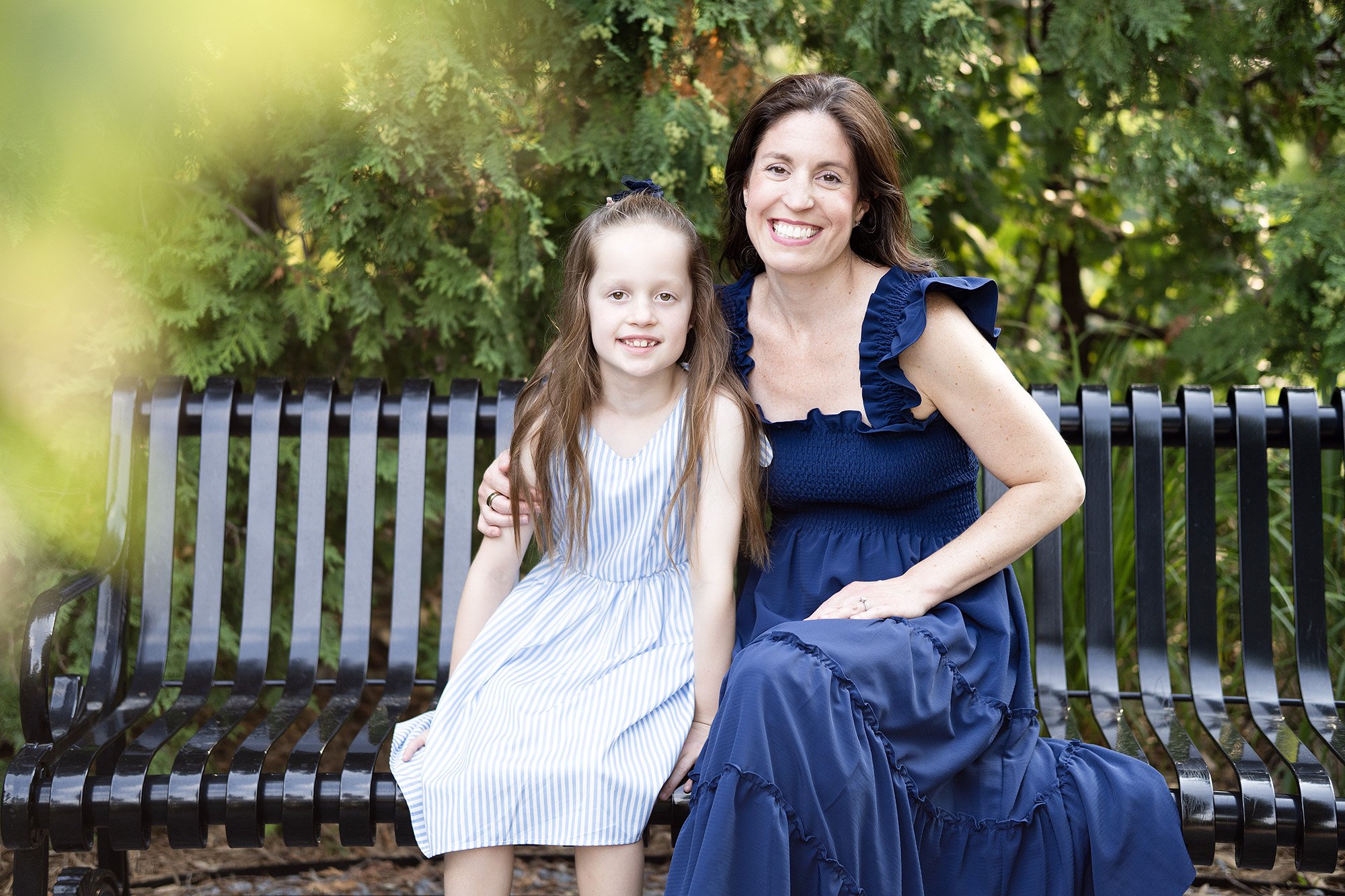 A woman and a young girl sitting on a black park bench surrounded by green trees. The woman is smiling, wearing a blue dress with ruffled sleeves, and has dark wavy hair. The girl, also smiling, has long light brown hair and is wearing a light blue a