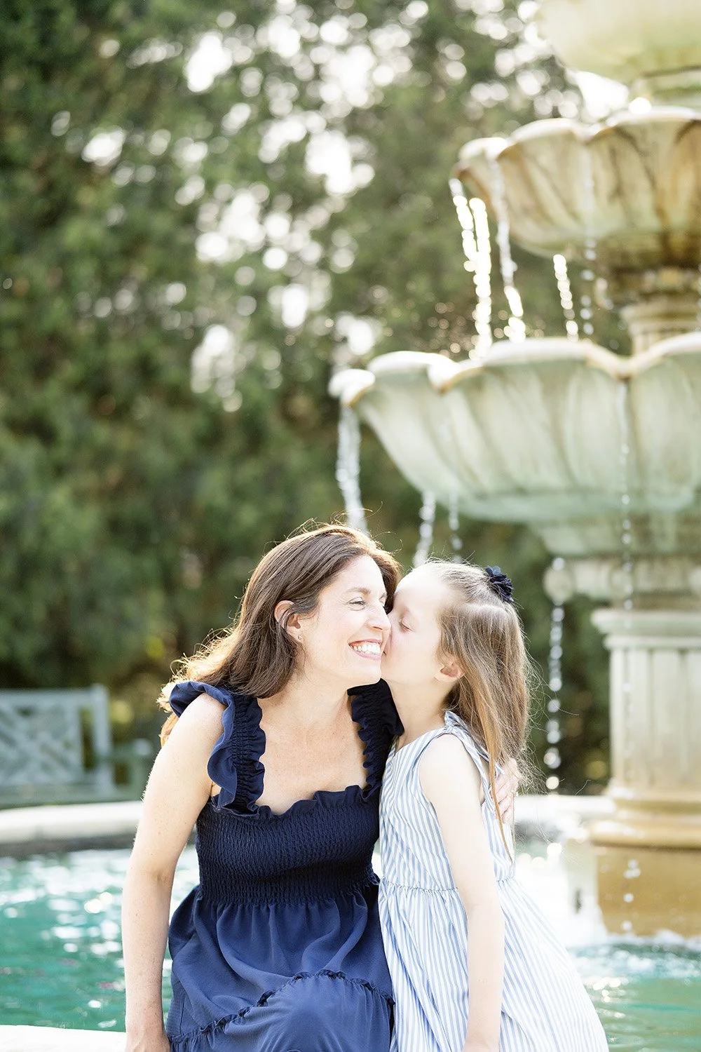 A woman and a young girl sharing a kiss by a fountain outdoors in a park, with trees in the background.