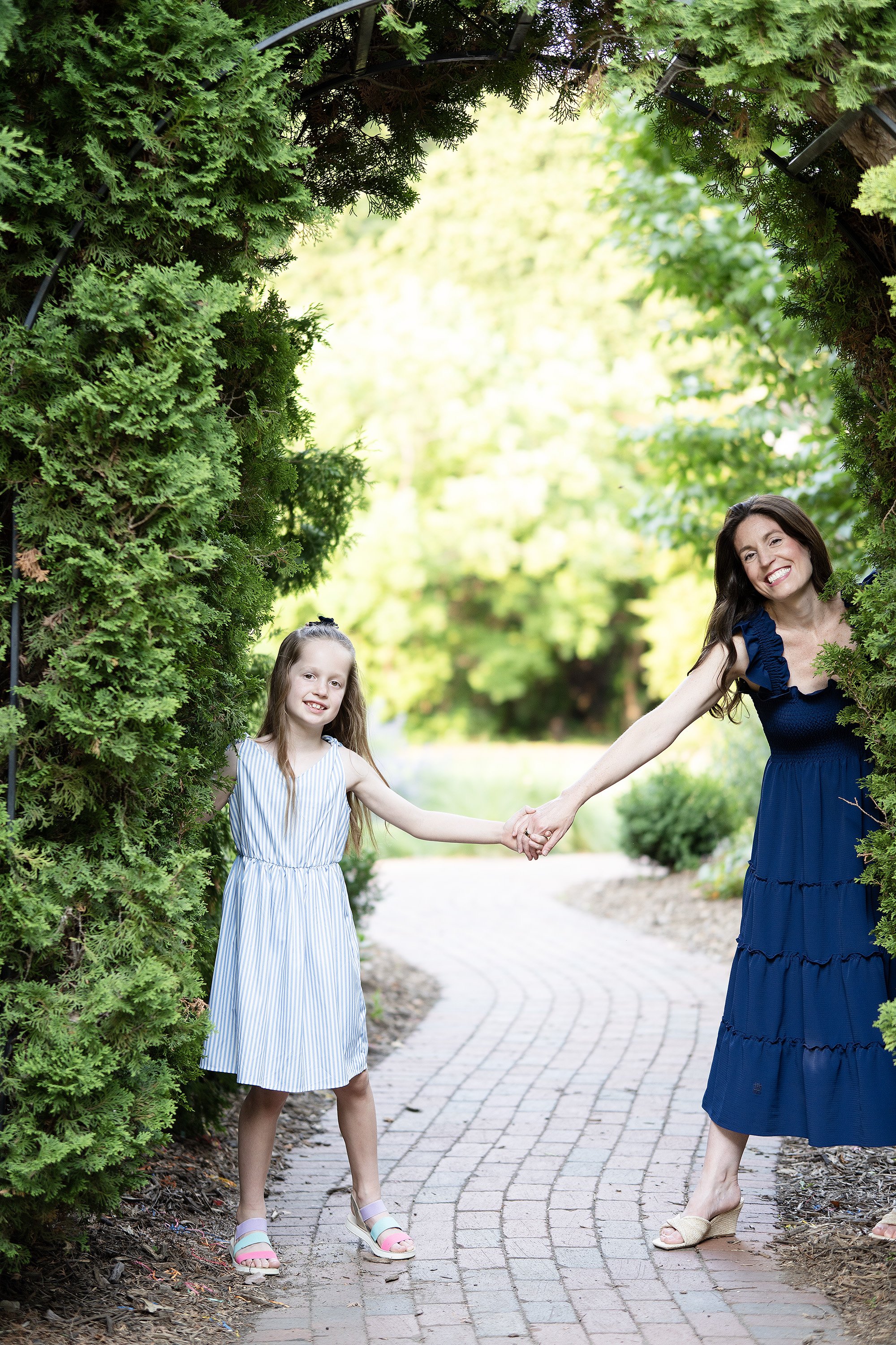 A woman and a girl are standing in a garden, holding hands and smiling. They are framed by green shrubbery on either side, with a brick pathway in the background.