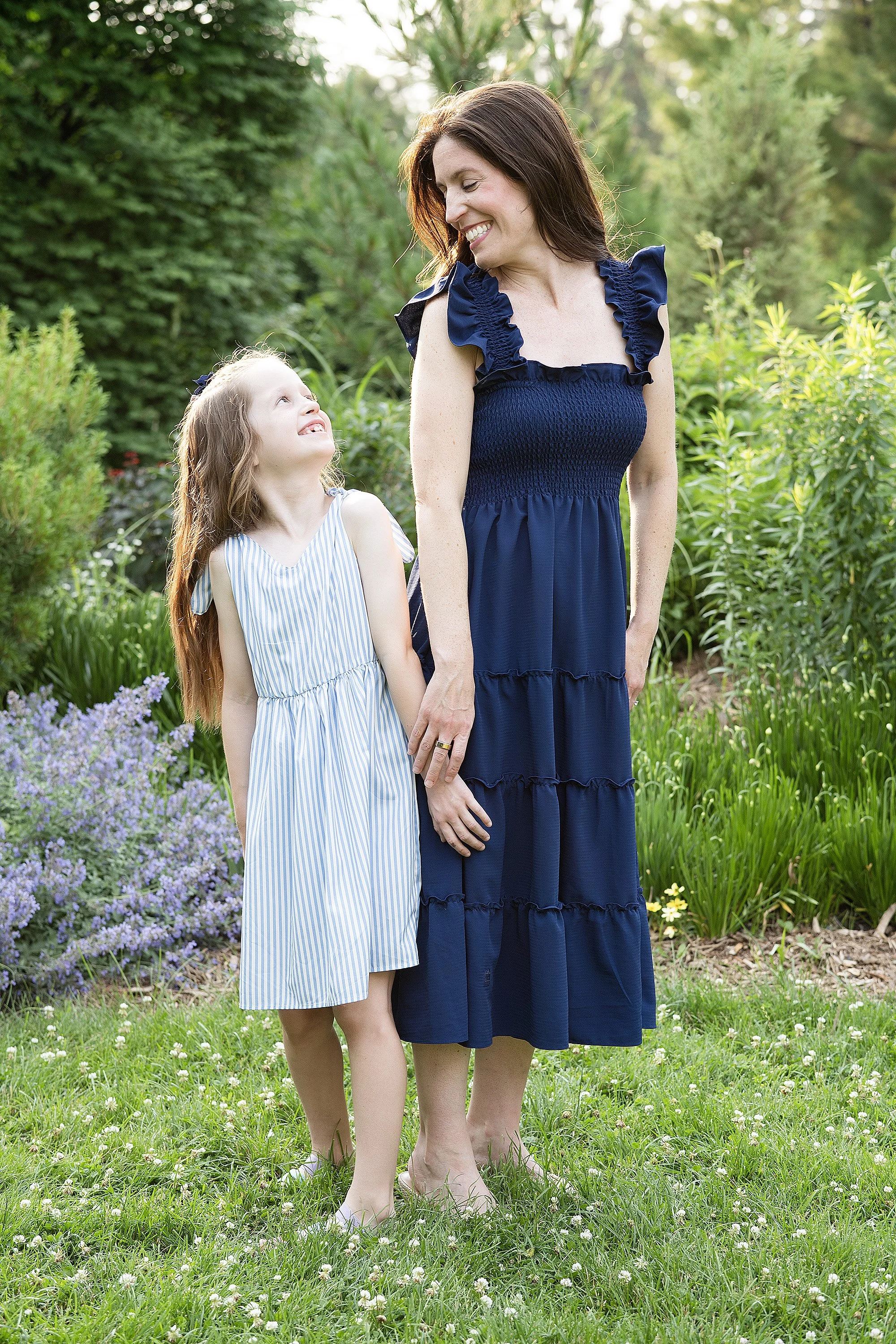 A woman and a young girl standing together outdoors in a garden with green foliage and purple flowers. The woman is smiling and looking at the girl, who is looking up at her with a smile.