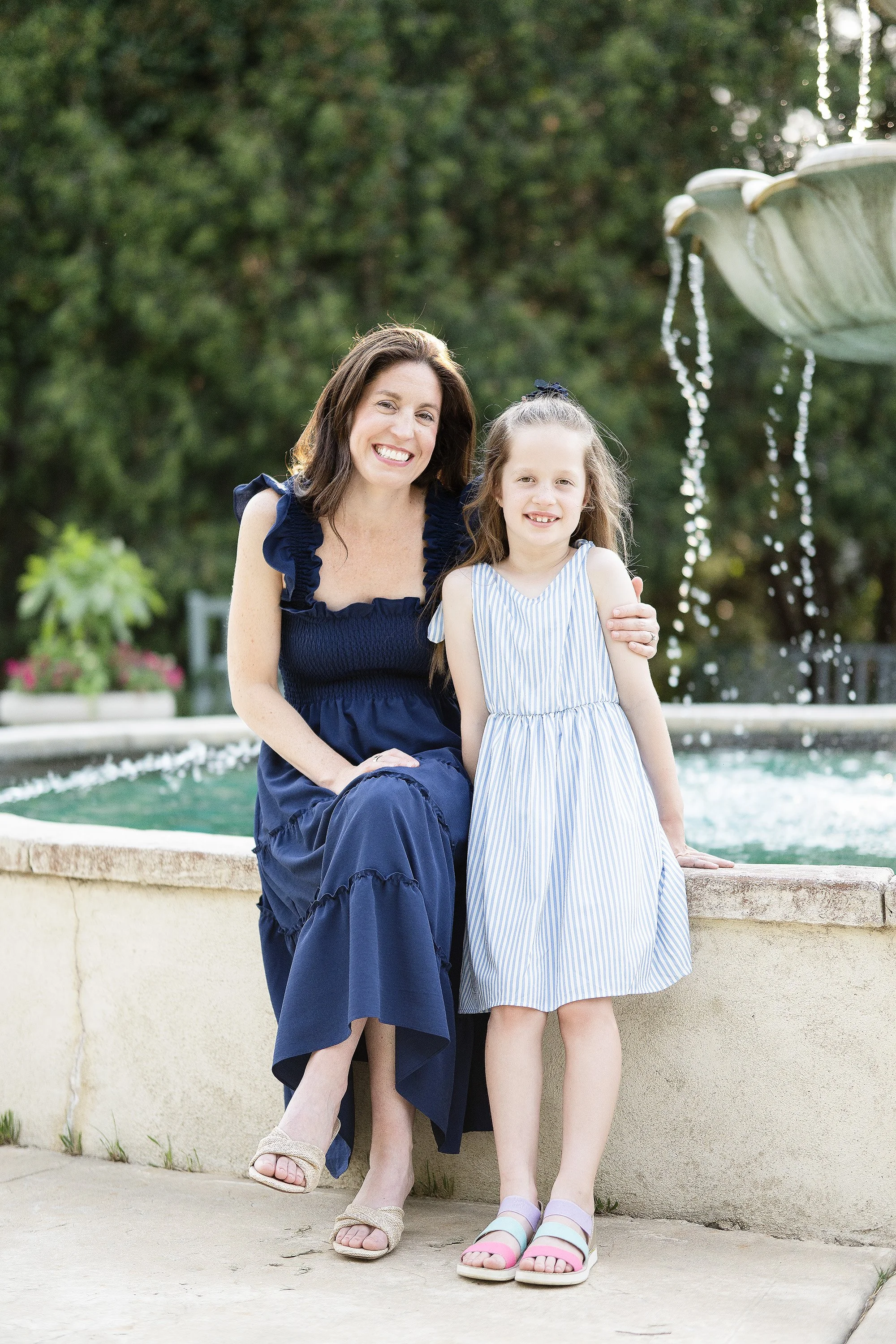 A woman and a girl sitting at a fountain outside, smiling. The woman has dark hair and is wearing a navy blue dress, and the girl has light hair tied with a bow, wearing a blue and white striped dress.