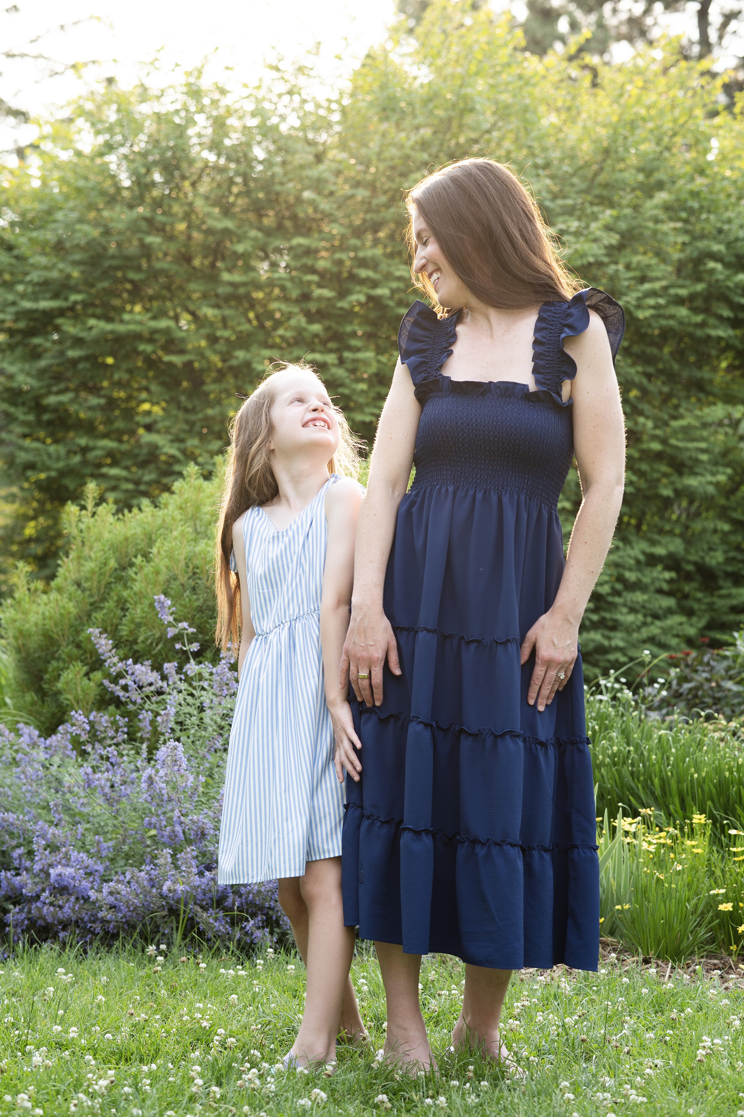 A woman in a navy blue dress smiling and looking at a young girl in a striped dress, standing in a garden with green trees and purple flowers, during daylight.