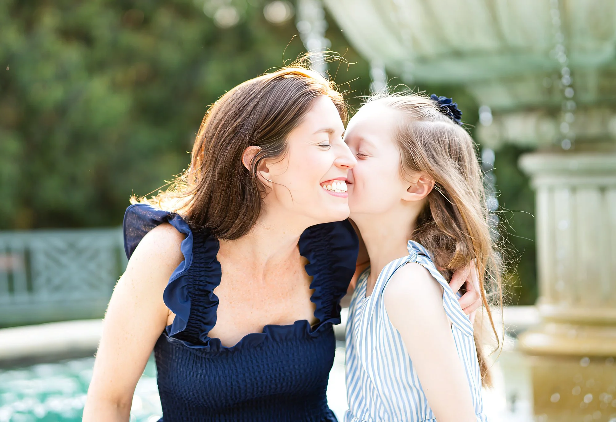 A woman and a young girl with long hair sharing a close and joyful moment outdoors during daylight, with the woman smiling and the girl about to kiss her cheek.