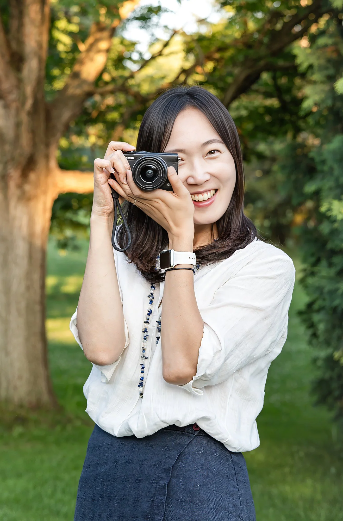 A woman smiling and holding a camera up to her face outdoors in a park with trees and greenery in the background.